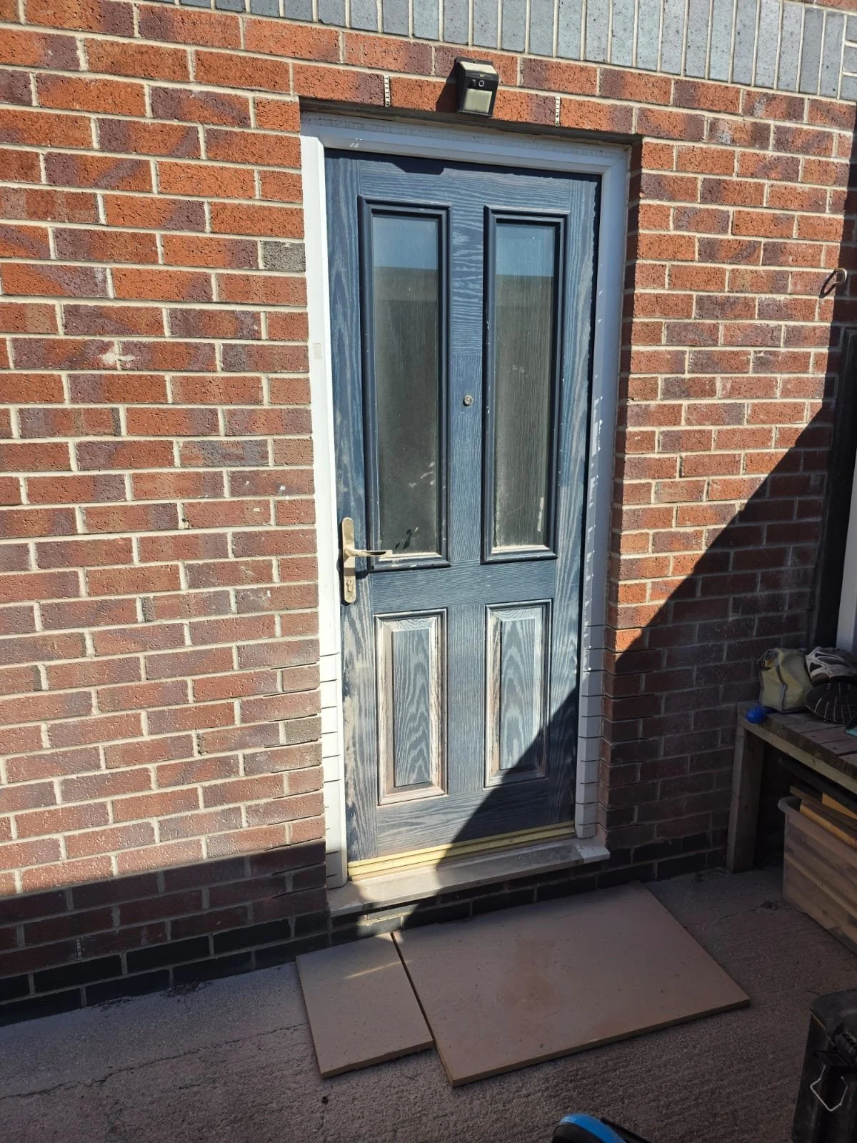 A dark blue door with glass panels is set into a red brick wall. The door appears to be newly painted, with some paint smudges visible. A small outdoor light is mounted above the door, and there are tiles and construction tools on the ground nearby, 