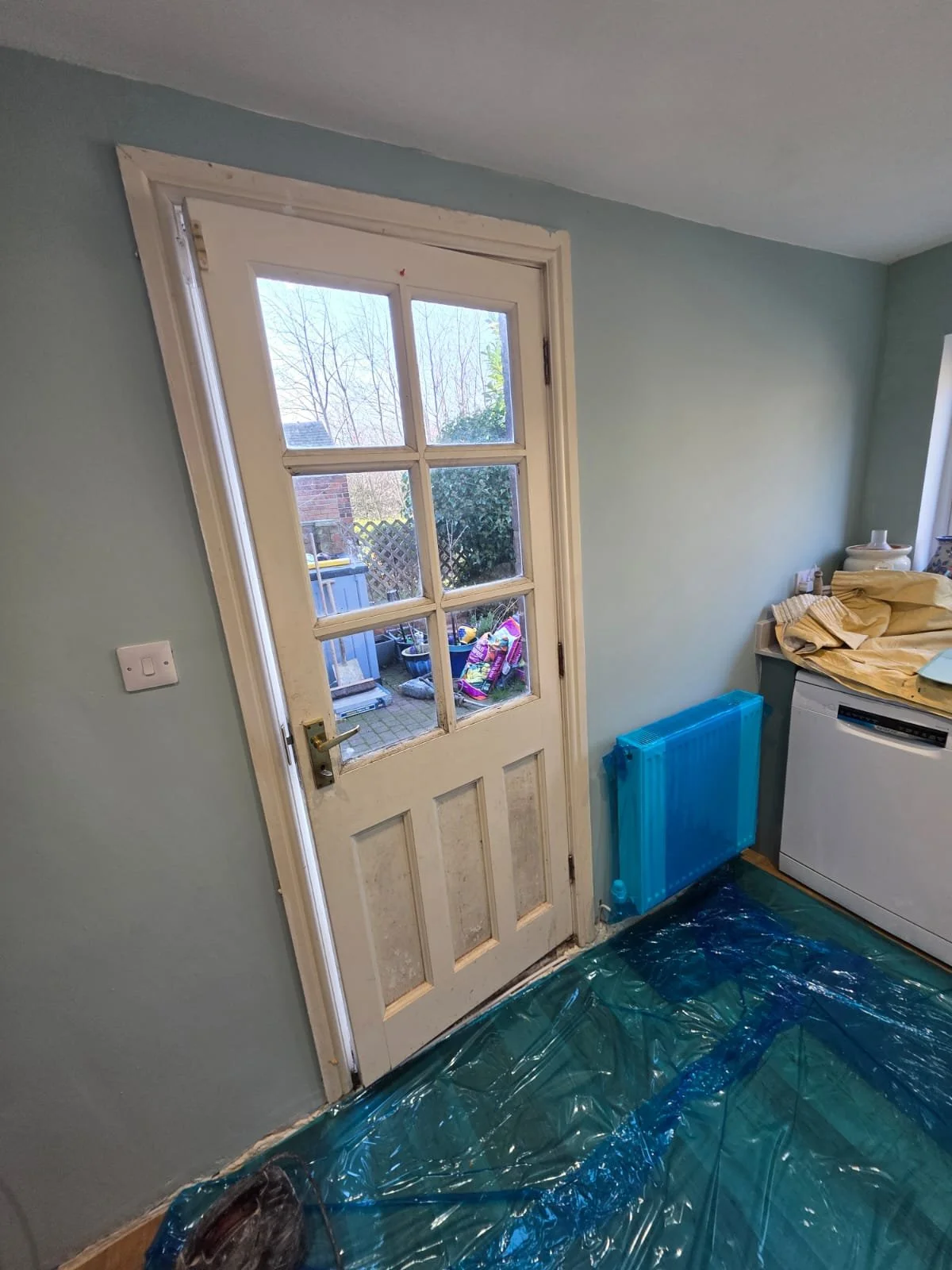 Room with a white door with glass panes, a blue radiator, and a washing machine. There is a table with cloth and various items on it. The floor is covered with a green plastic sheet.