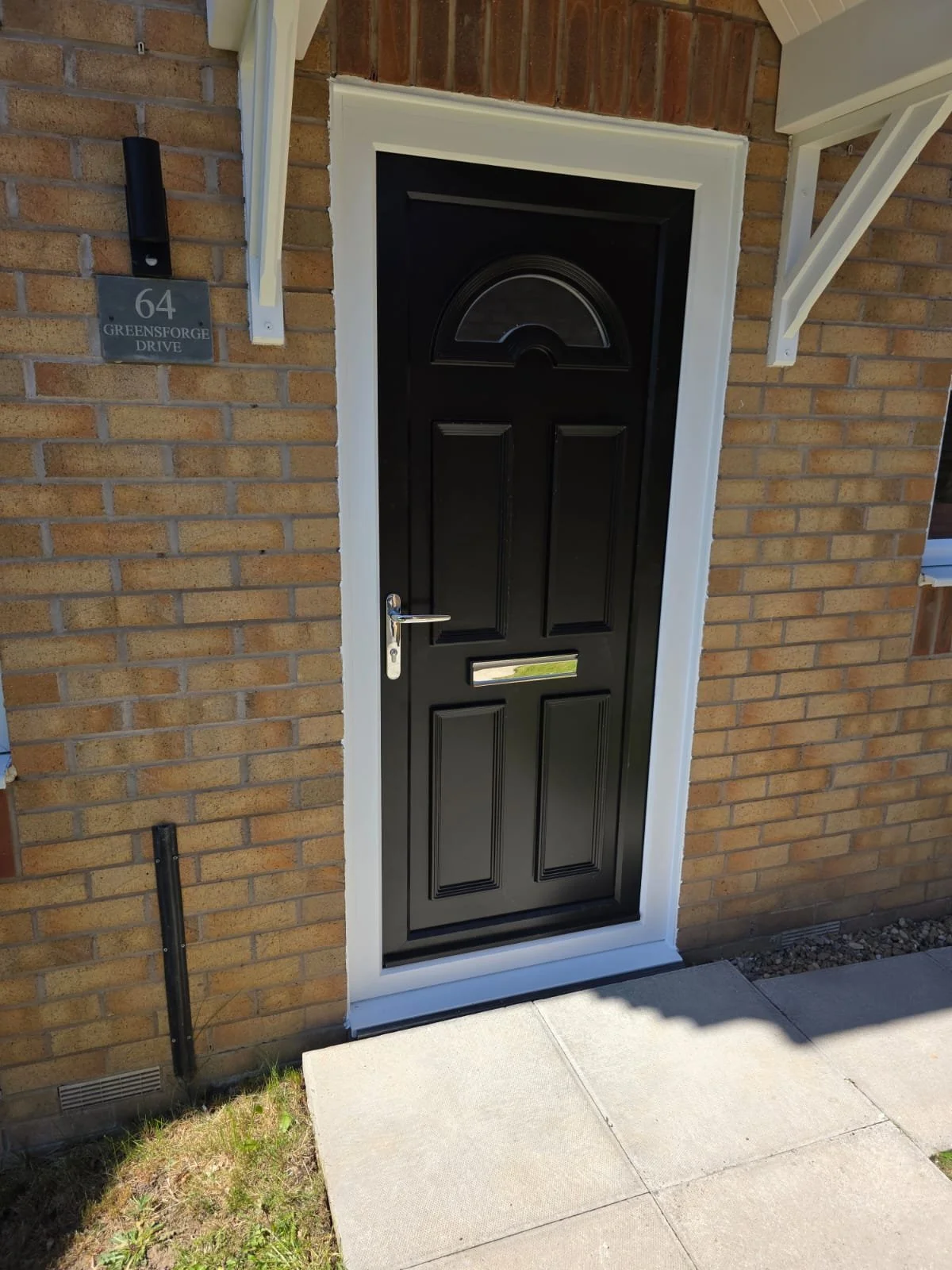 Black front door with a small, arched window at the top, set in a brick wall with white trim. The door has a silver handle and mail slot. To the left of the door is a house number sign reading '64 Greensforge Drive' and a black wall-mounted light. Th