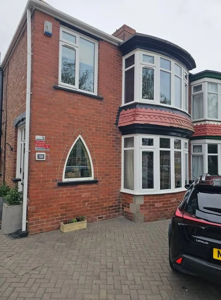 A two-story brick house with a front bay window, a small arched window, and a driveway with a black Lexus car parked. The house has white window frames and a red-tiled awning over the bay window.