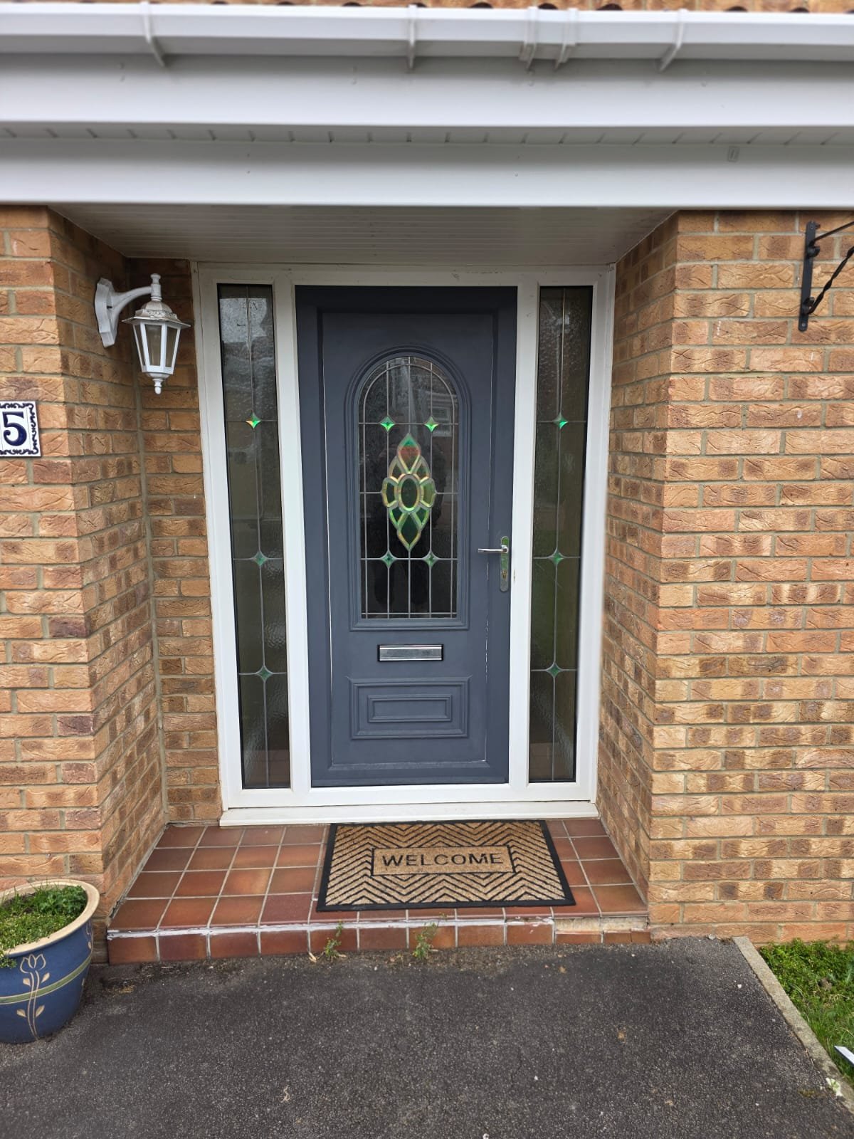 Front entrance of a house with a dark gray door, decorative glass panels, a welcome mat, brick walls on either side, a white outdoor light, and potted plants.