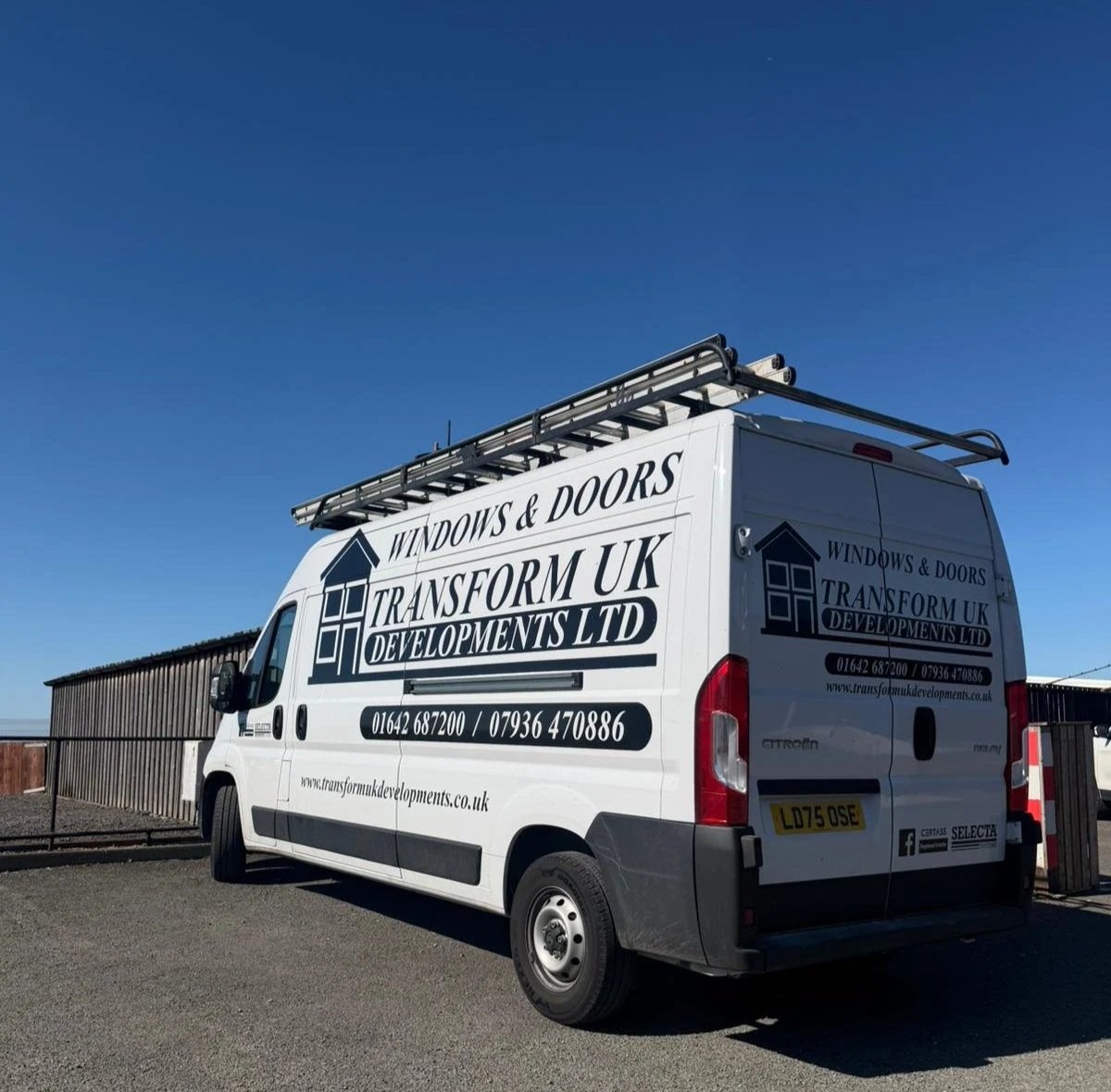 A white work van with the logo and contact information for Windows & Doors Transform UK Developments LTD parked outdoors under a clear blue sky.