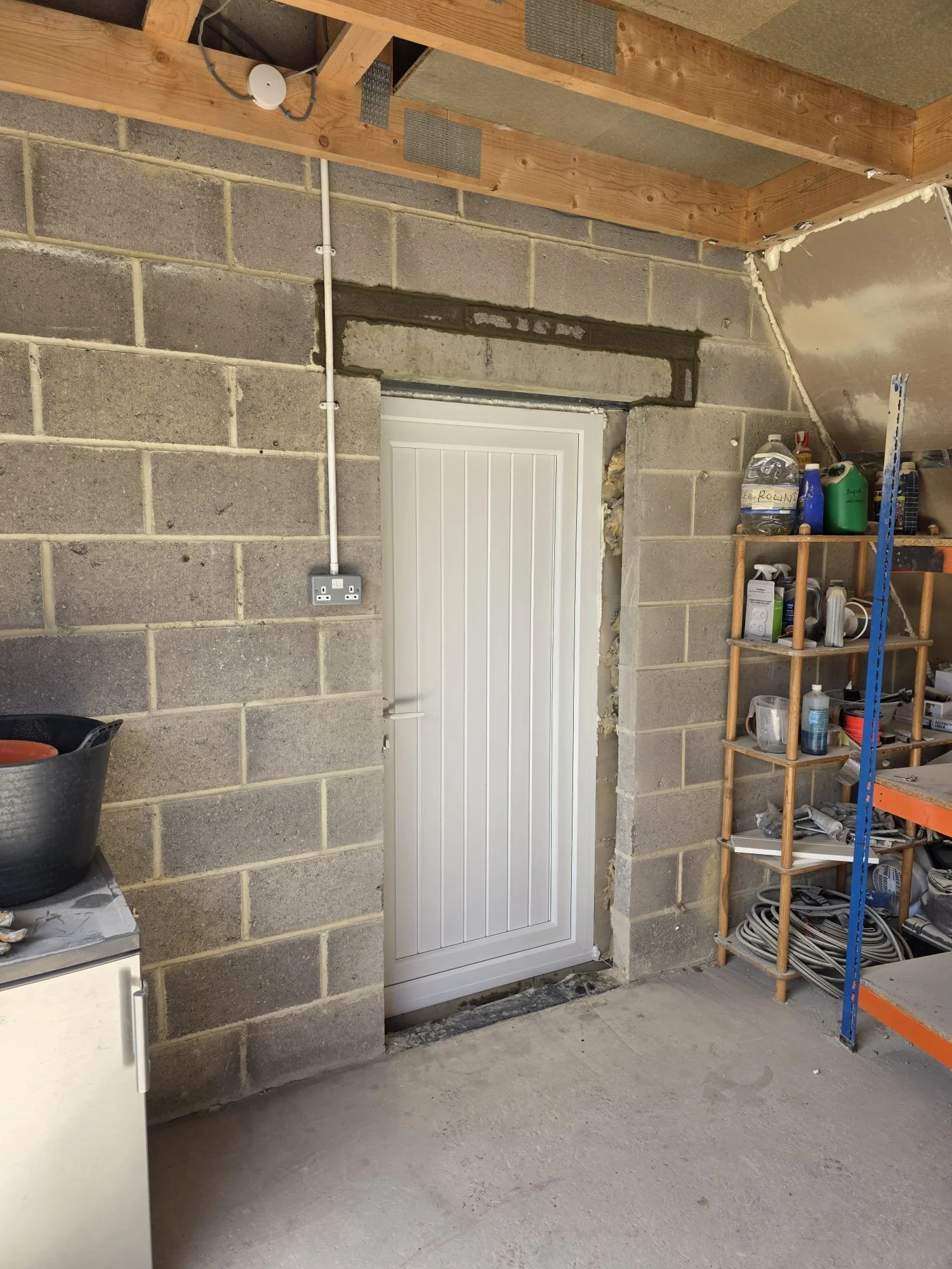 Unfinished interior wall with cinder blocks, electrical wiring, a white door, andwork shelves with various cleaning supplies and tools.