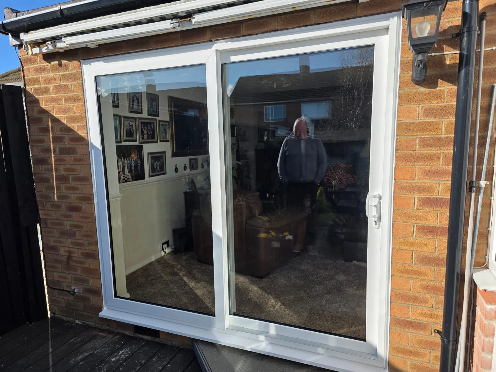 A sliding glass door on a brick house showing the reflection of a man inside a room with multiple framed pictures on the wall.