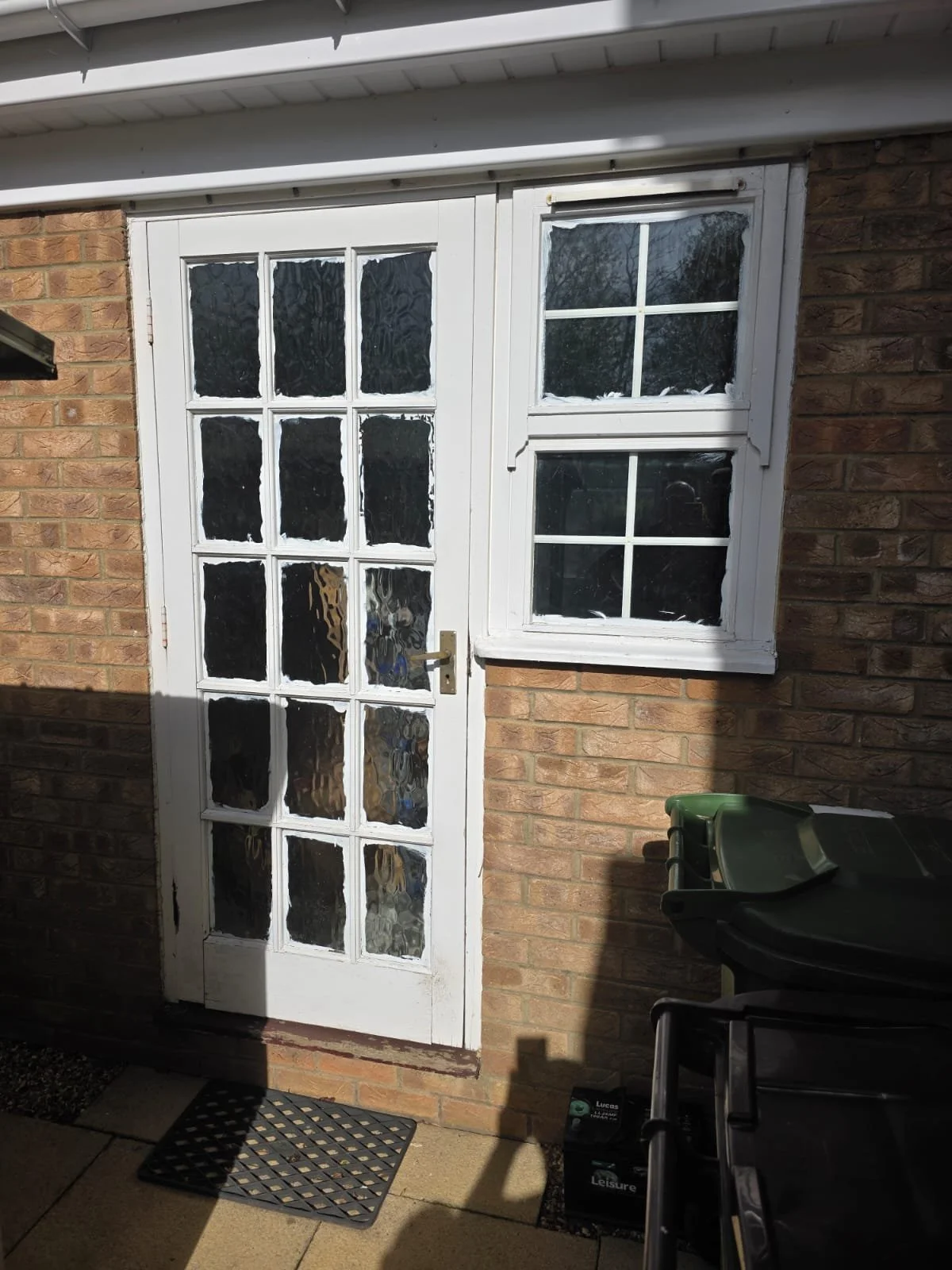 A white door with glass panes next to a white-framed window with diamond-shaped muntins on a brick wall exterior. A black mat is on the ground in front of the door. Shadows are cast on the wall and ground. A green trash bin and gardening tools are vi