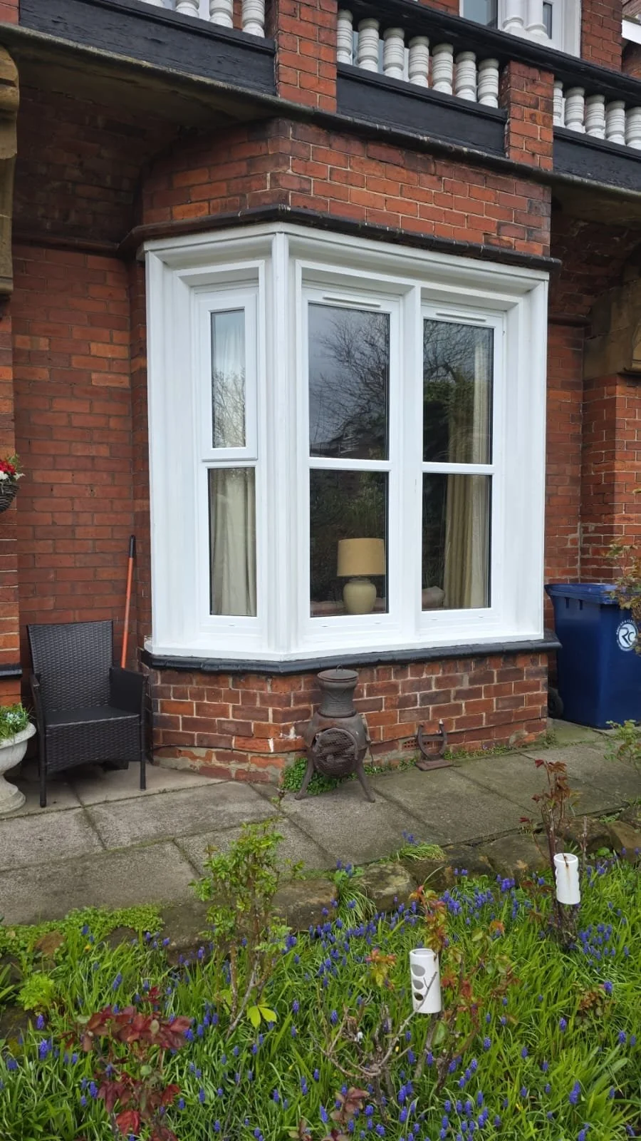 Red brick house with a large white bay window, some backyard furniture including a black chair, garden plants with purple flowers, a blue trash bin, and old metal wood stove