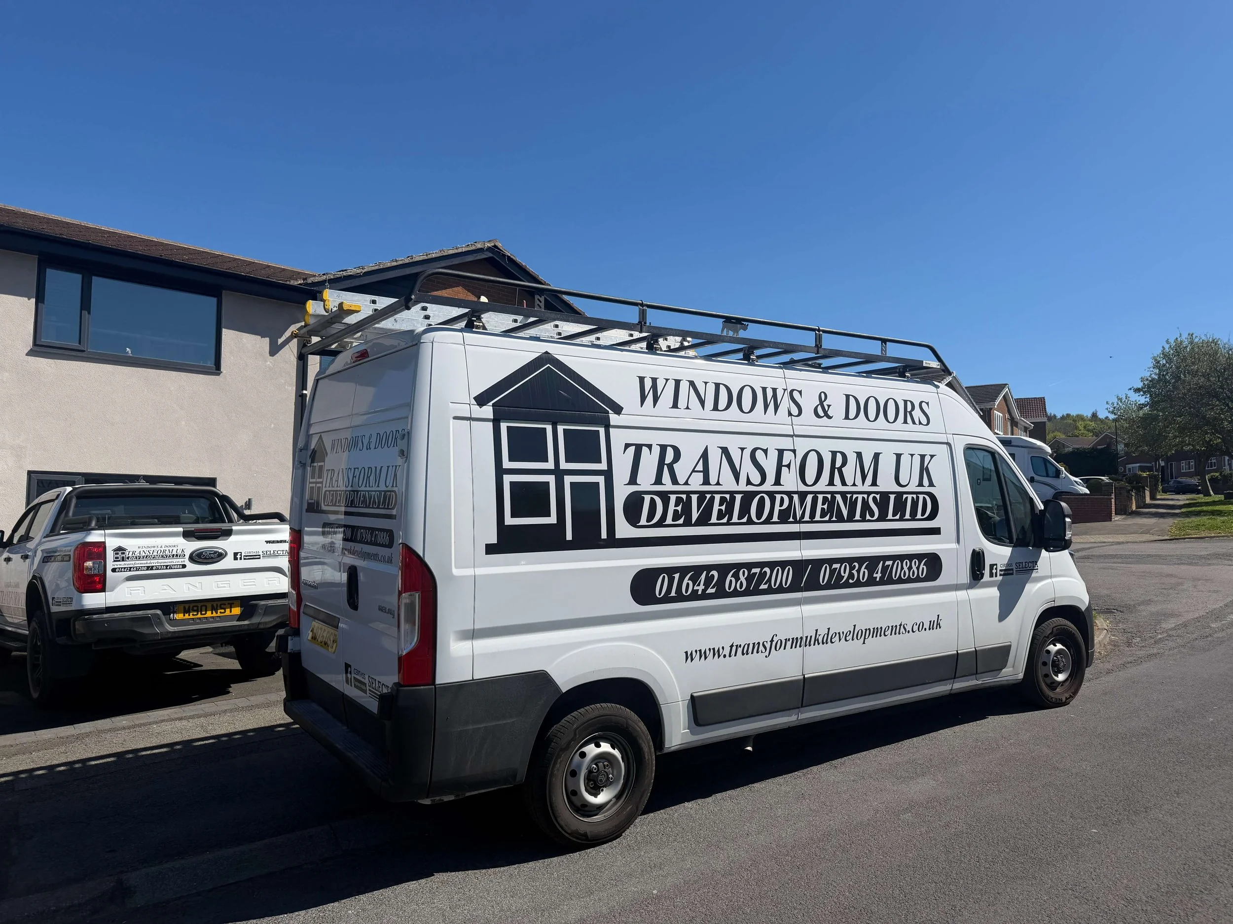 A white van with black and yellow logo and text for Transform UK Windows & Doors, parked on a residential street under a clear blue sky. The van has a roof rack with tools. In the background, houses with front gardens and a second vehicle are visible.