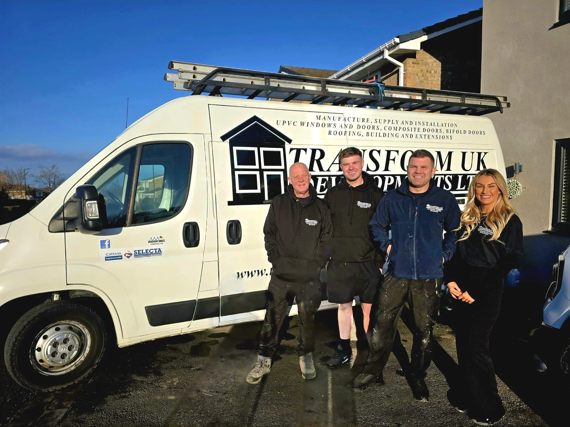 A white work van parked outdoors with a ladder on top, and four people standing in front of it, all smiling. The van has company information on the side. The background includes a residential building with a clear blue sky.