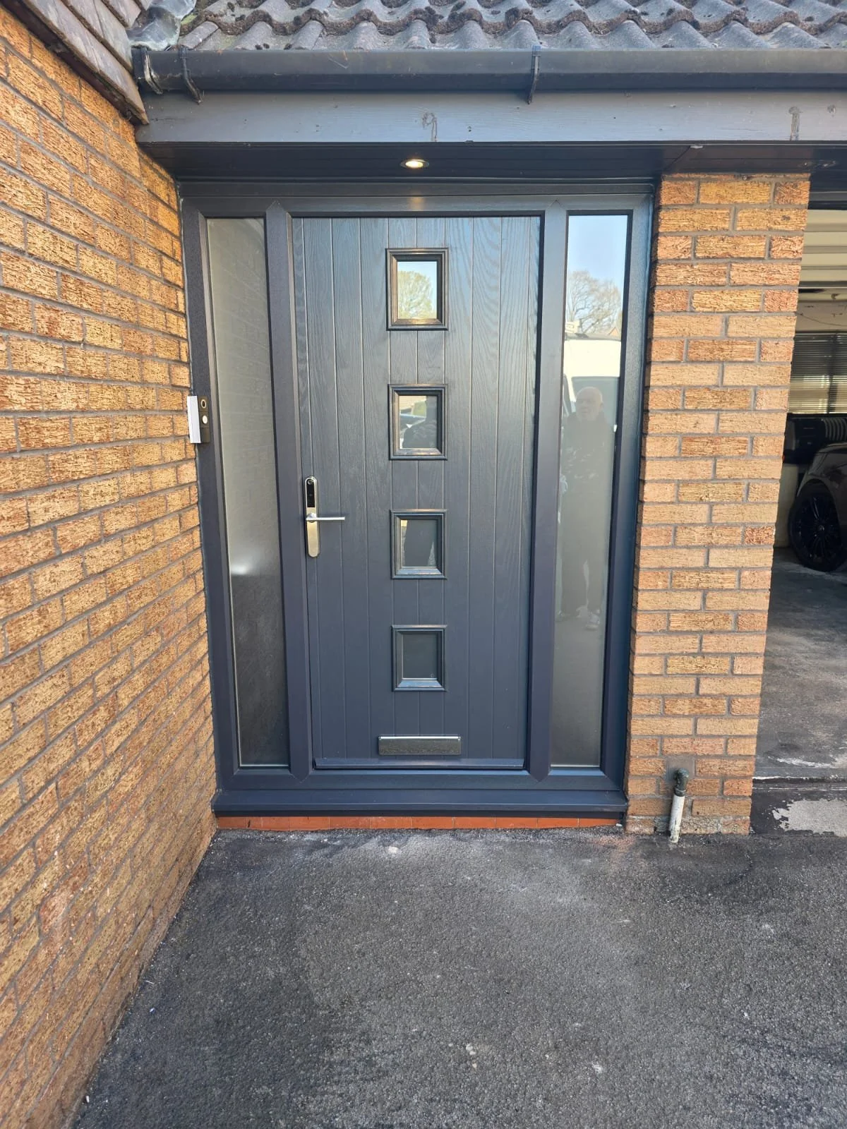 A front door made of dark colored wood with four small glass panes, a silver handle, and a mail slot, set within a brick wall of a house.