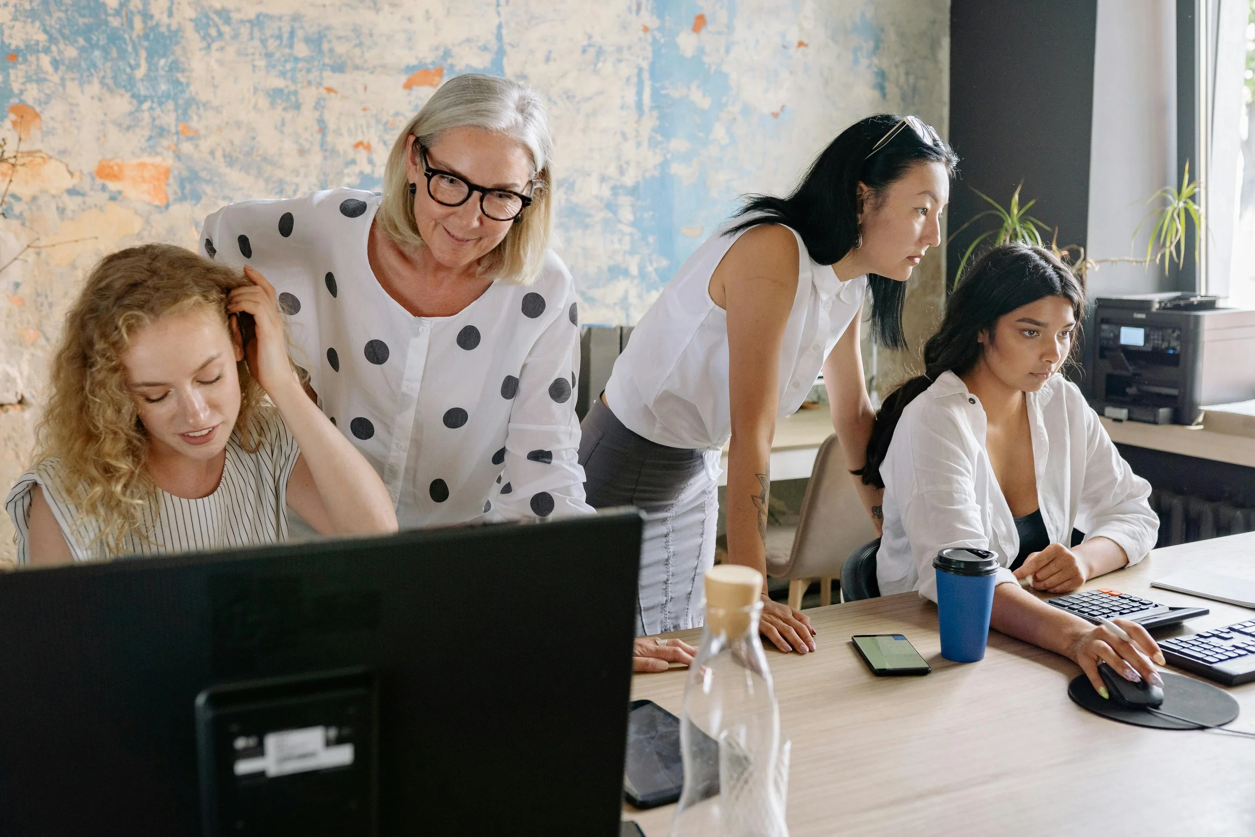A group of Asian and Caucasian women working together in an office. One woman with glasses and blonde hair is standing and leaning over, while others sit at a desk and focus on their tasks.