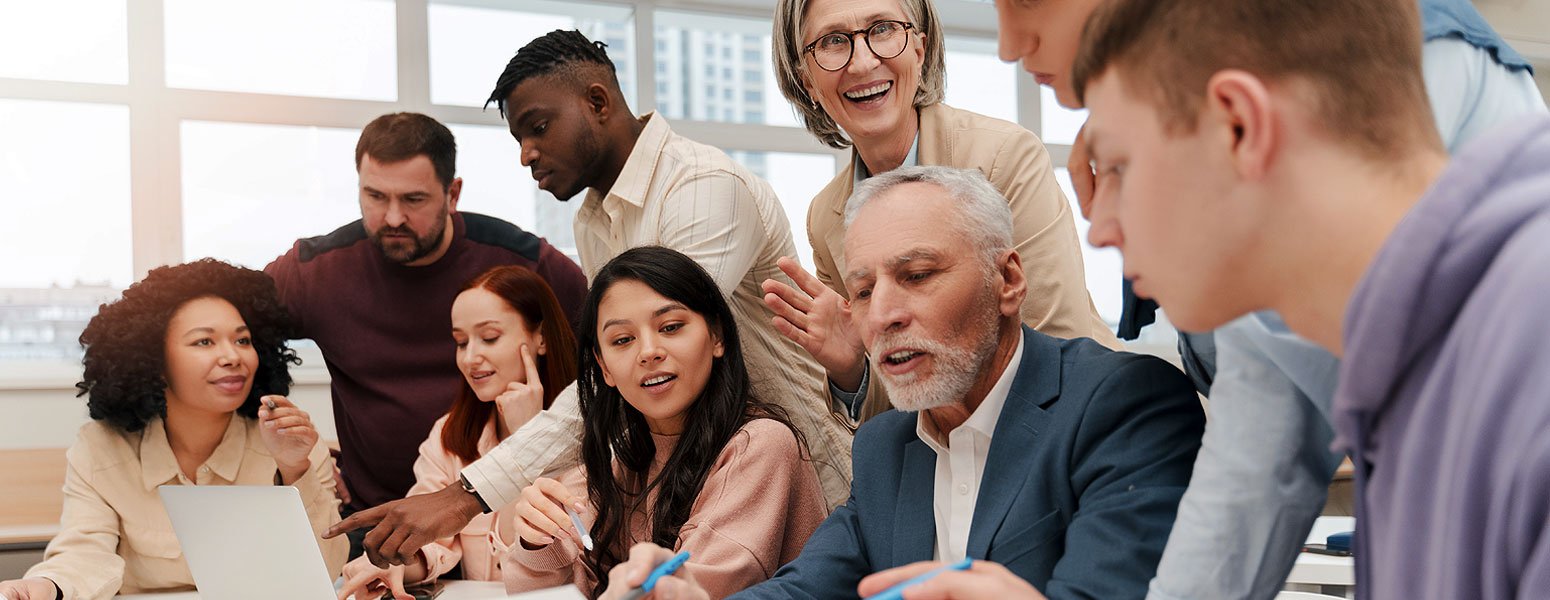 Group of diverse professionals, including men and women of different ages, engaged in a discussion around a table in a bright office, with large windows and city buildings in the background.