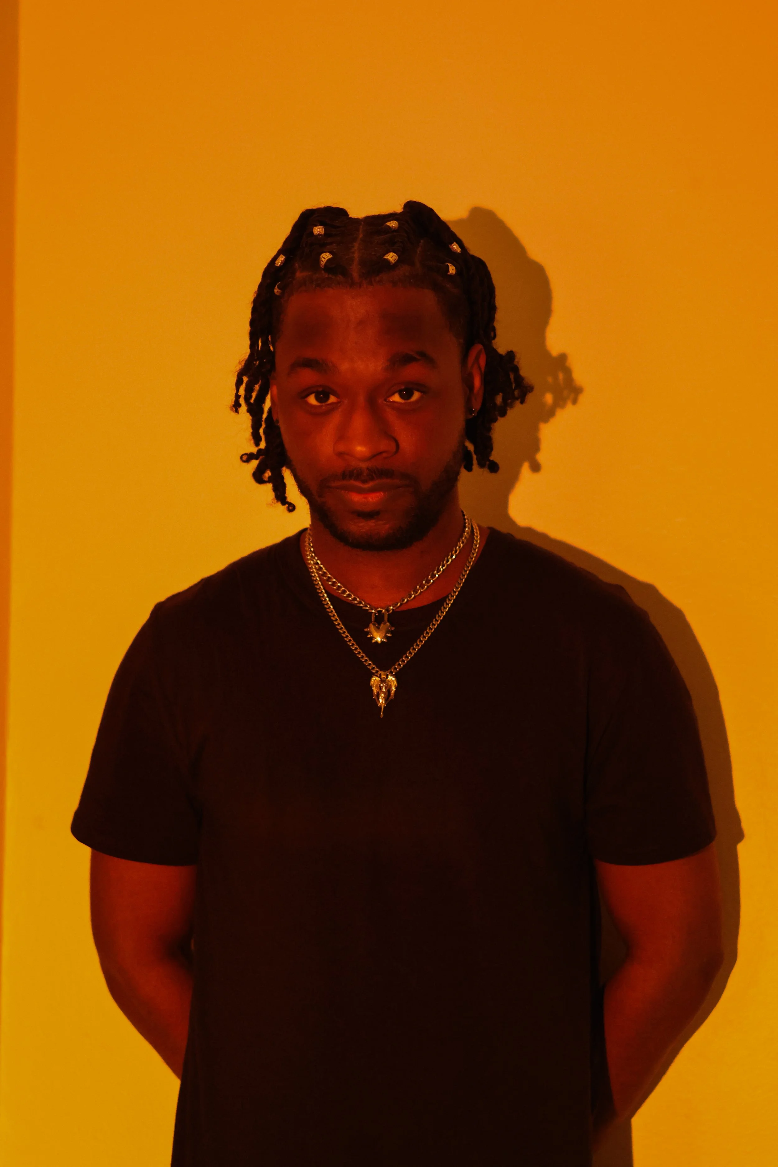 A young man with dreadlocks decorated with beads, wearing a black shirt and layered gold necklaces, stands against a plain yellow wall, casting a shadow to his right.