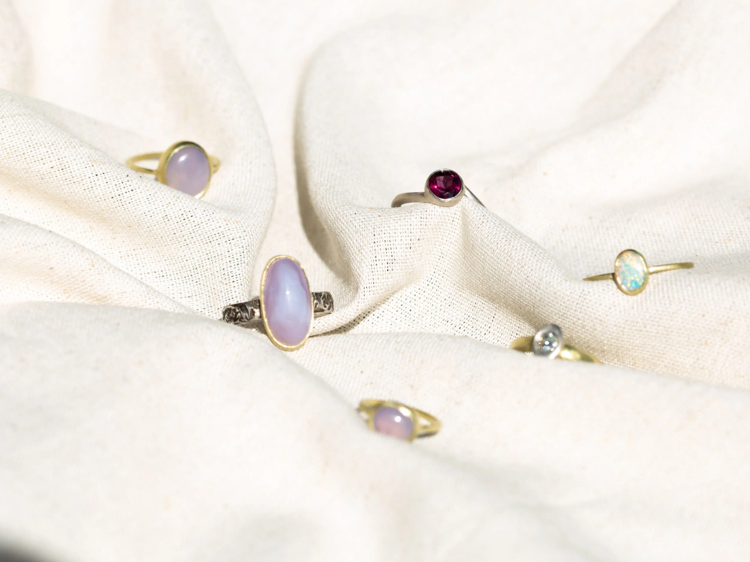 Several rings with gemstone settings, including a ring with a large Holley Blue Chalcedony cabochon, displayed on cream-colored fabric.