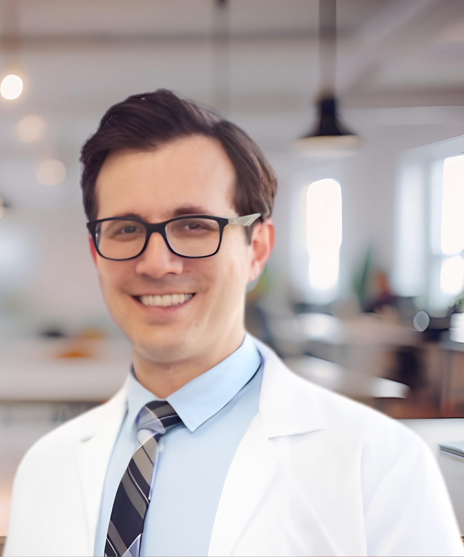 A smiling man wearing glasses, a white lab coat, a light blue shirt, and a striped tie, standing in a modern, well-lit kitchen.