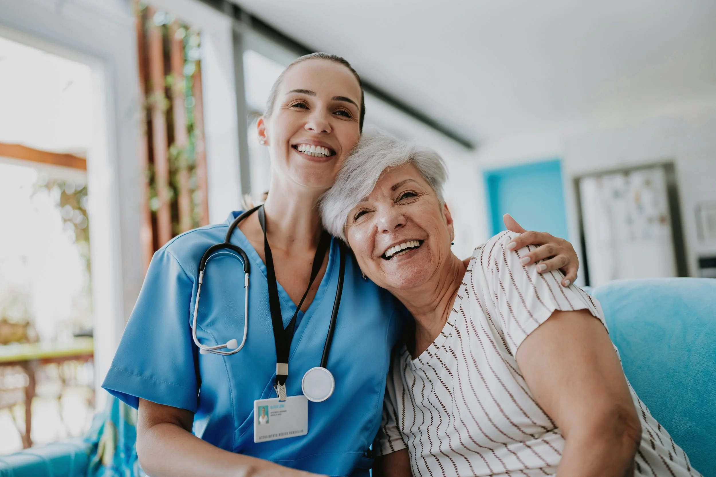 A female healthcare worker in blue scrubs and a stethoscope hugging an elderly woman with gray hair, wearing a striped shirt, in a bright clinical setting.