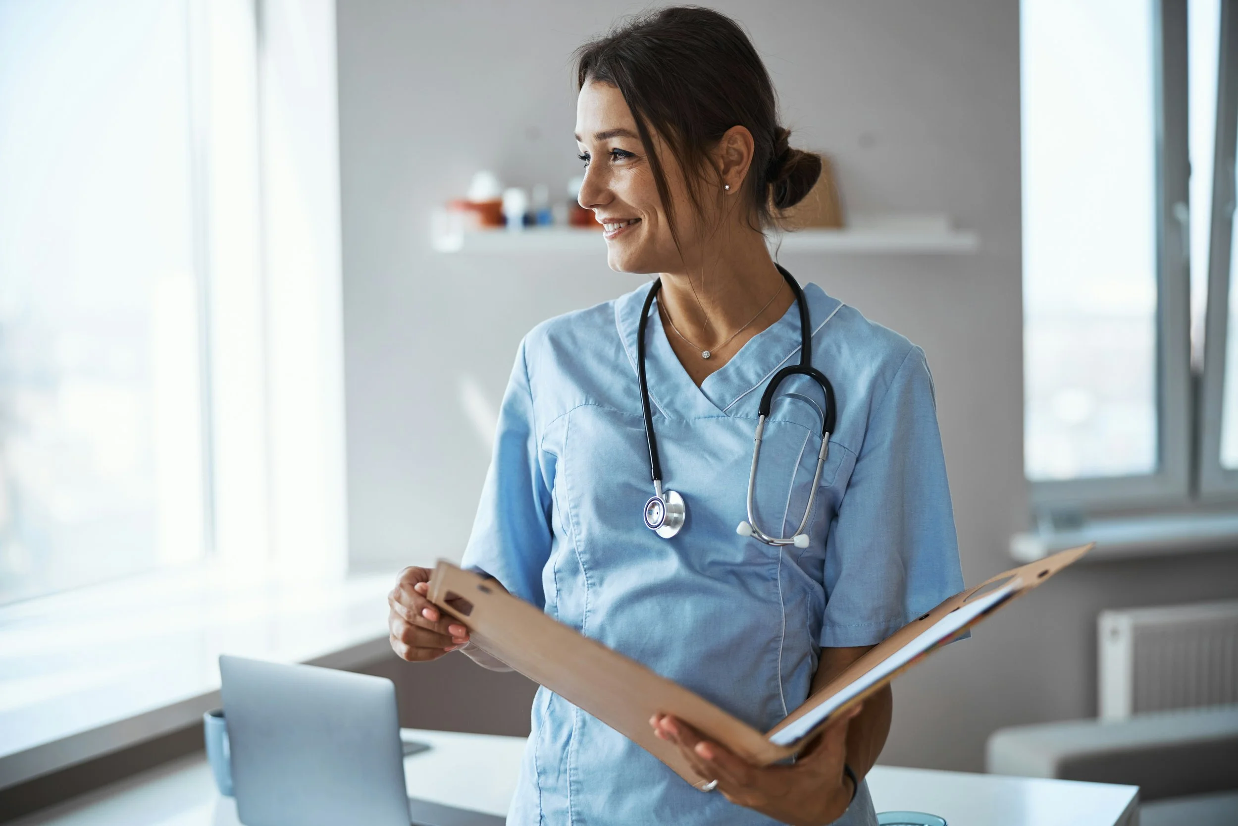 A female healthcare worker in light blue scrubs with a stethoscope around her neck, holding a clipboard and smiling in a bright medical office.