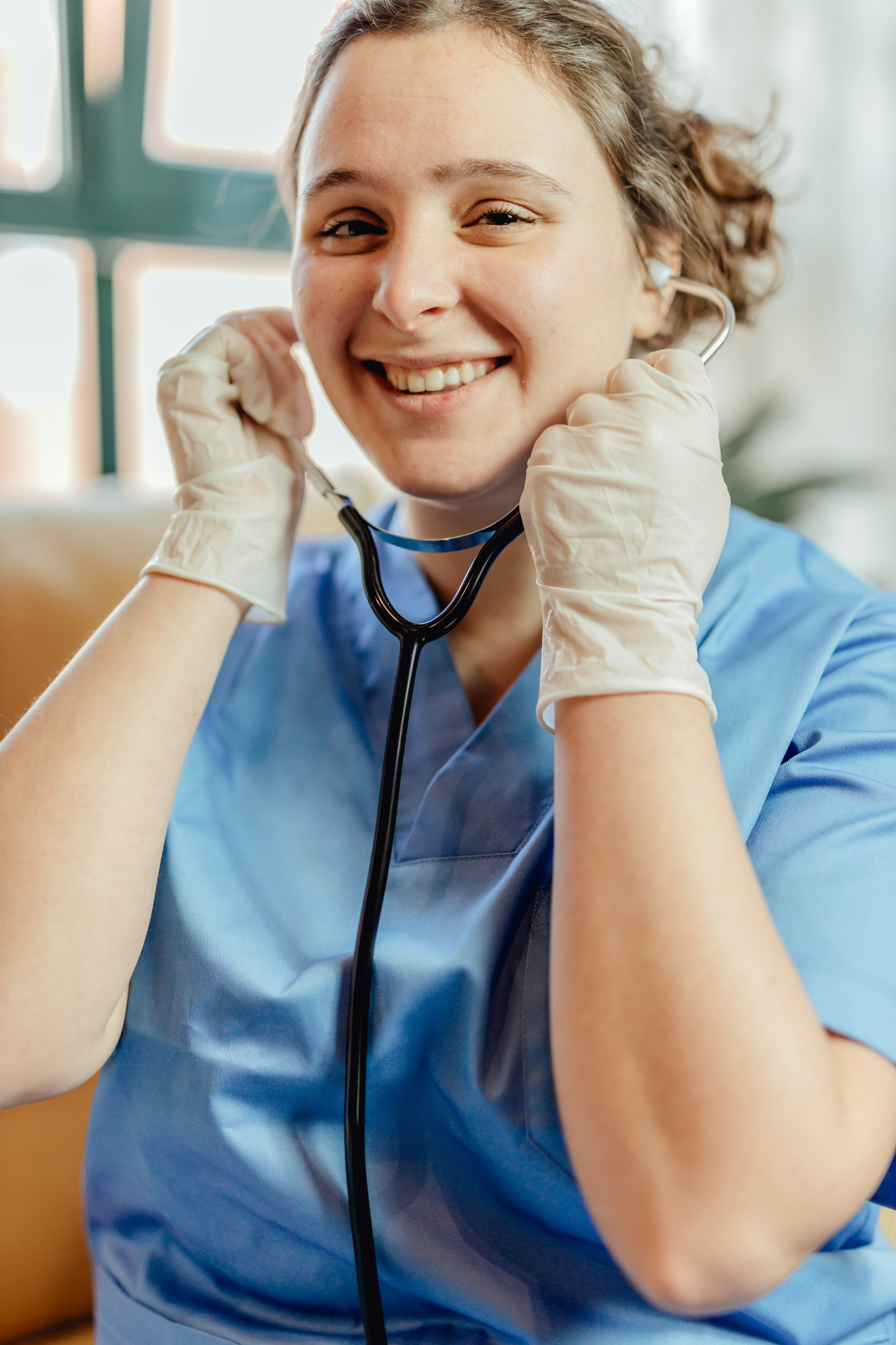 A young woman wearing medical scrubs and gloves is smiling and holding a stethoscope to her ear.