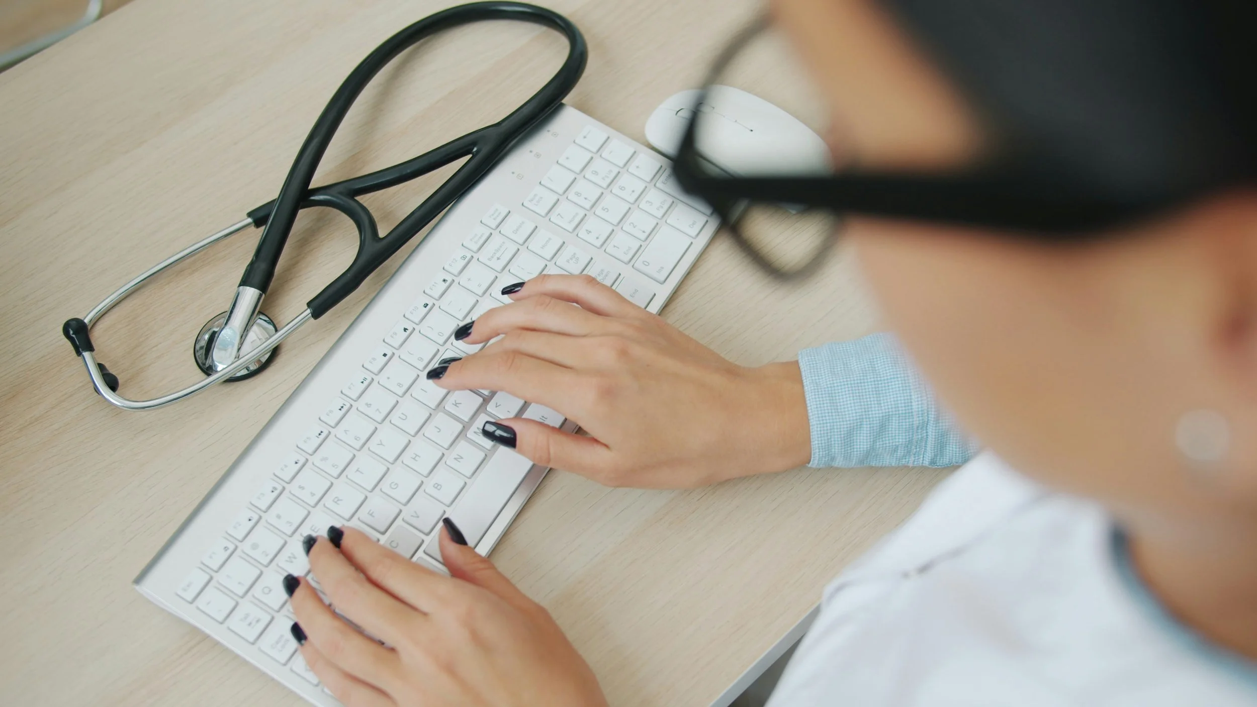 A person wearing glasses typing on a white computer keyboard at a desk, with a stethoscope placed nearby.
