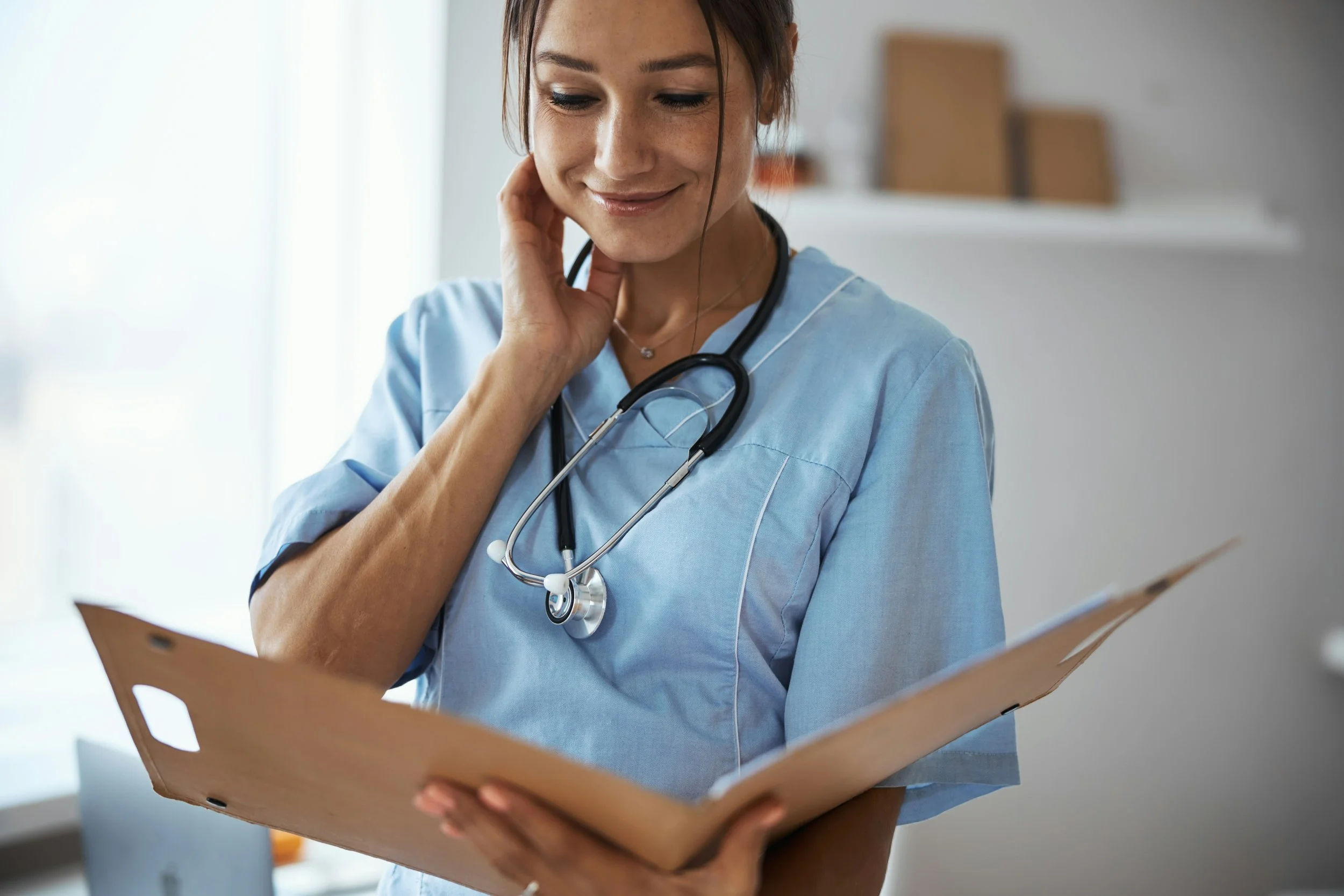 A female healthcare professional in light blue scrubs with a stethoscope around her neck reads a clipboard or medical chart, smiling gently.