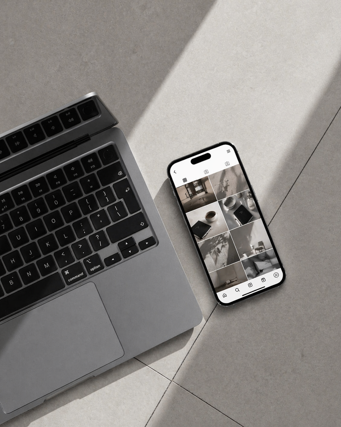 A silver laptop with a black keyboard and a smartphone showing a grid of photos, placed on a light-colored tiled surface.