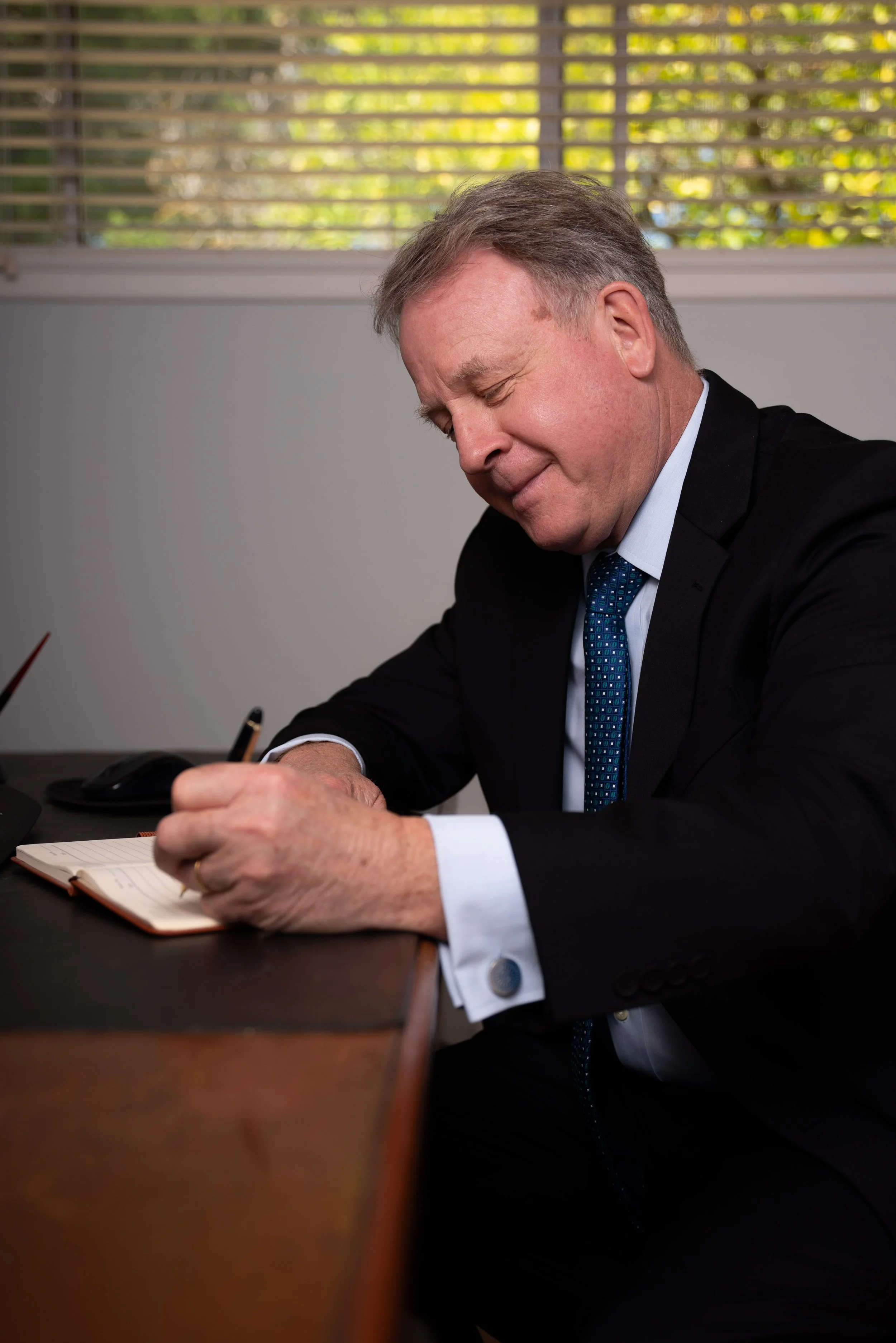 A middle-aged man in a business suit sitting at a desk writing in a notebook, with a window and blinds in the background.