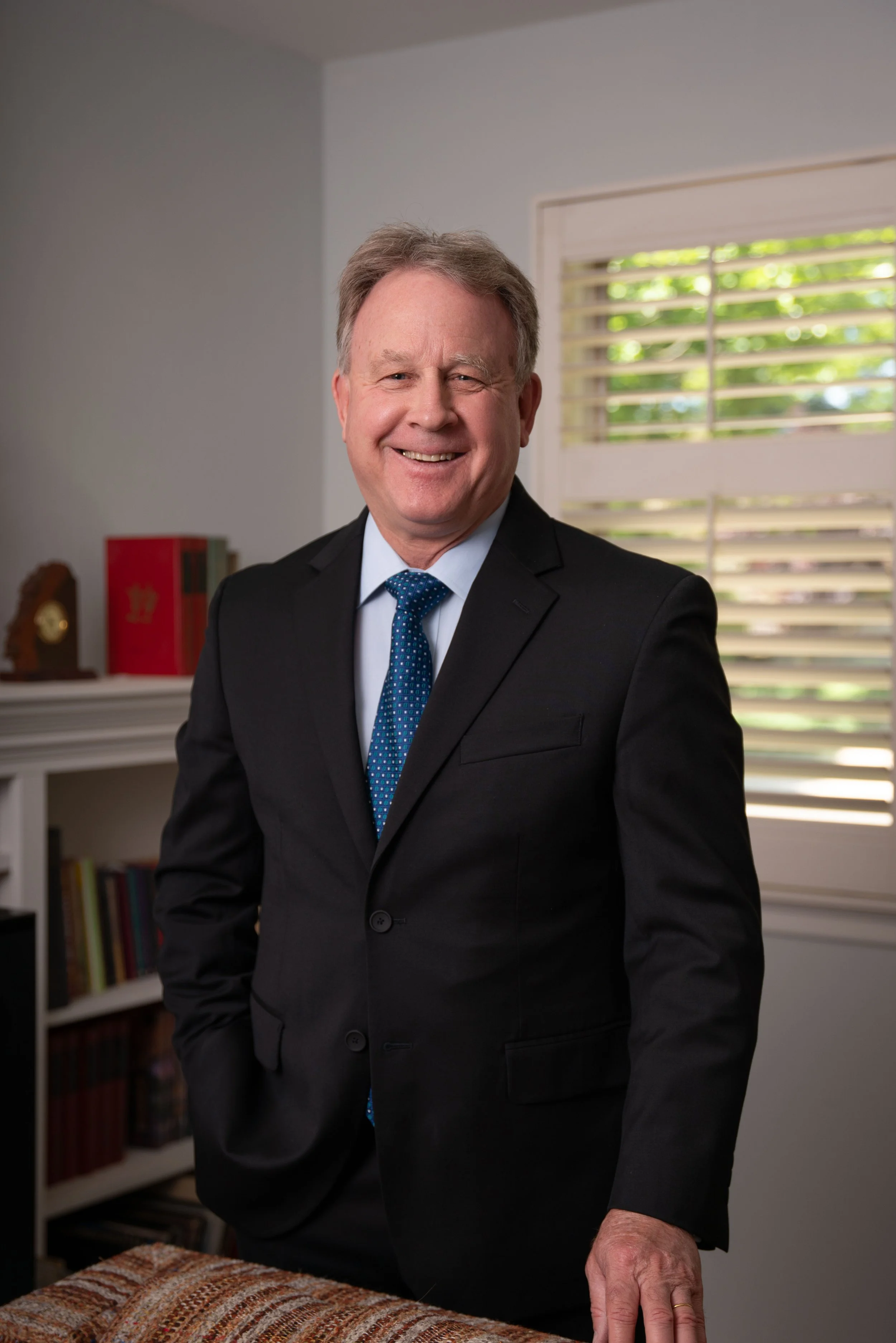 A smiling middle-aged man in a black suit, light blue shirt, and blue tie standing indoors in front of a window with blinds, with a bookshelf and a clock in the background.
