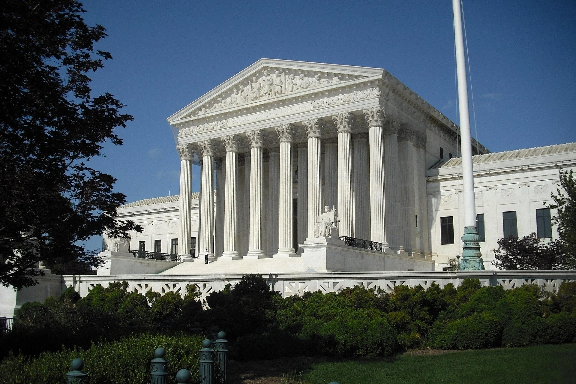 The United States Supreme Court building, a large white neoclassical structure with tall columns and statues, under a clear blue sky.