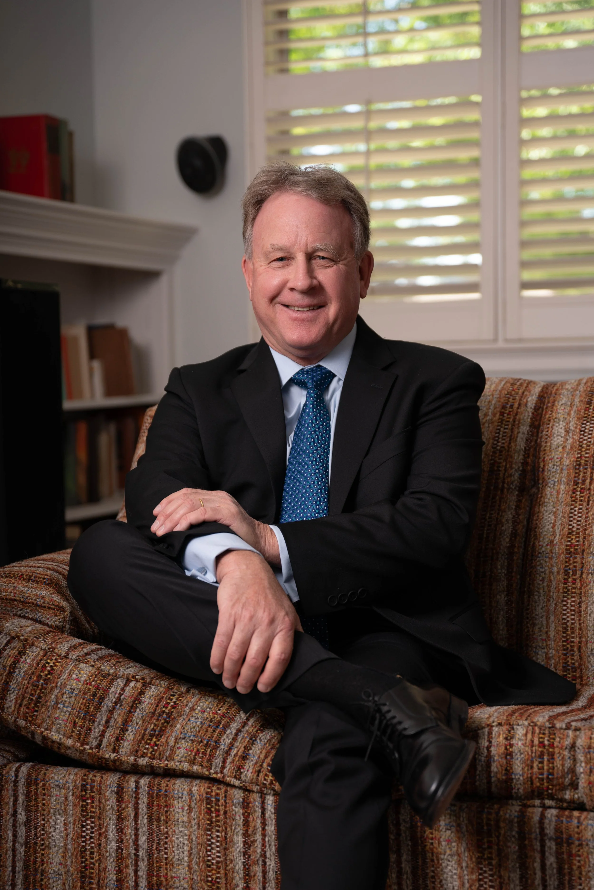A smiling middle-aged man sitting on a patterned sofa, wearing a black suit, white shirt, and blue tie, with a background of windows with blinds and a white wall with a bookshelf and a speaker.