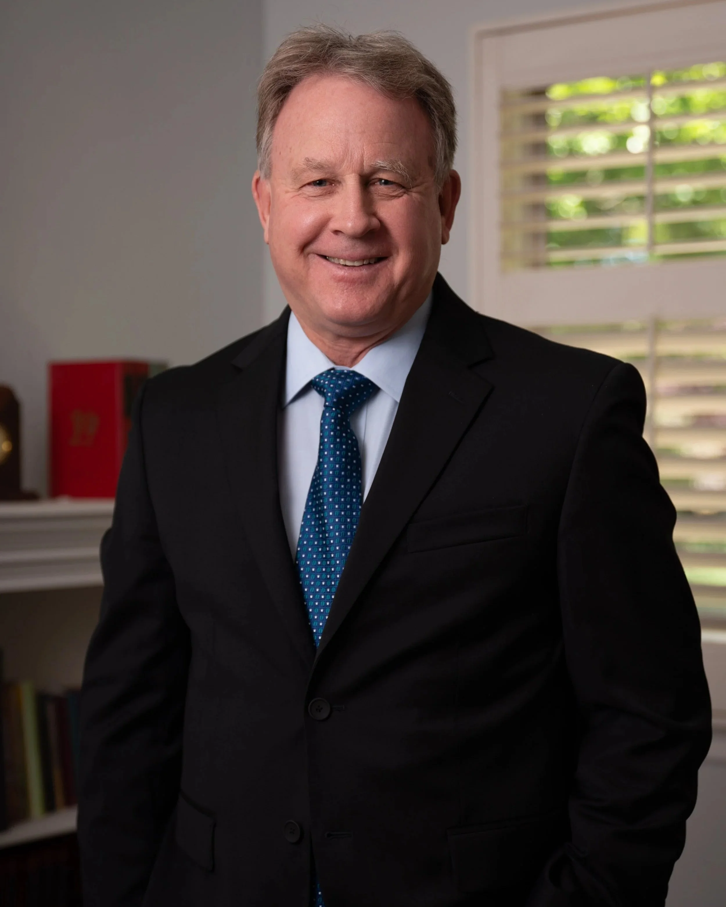 A middle-aged man with light brown hair wearing a dark suit, light blue shirt, and blue polka dot tie, smiling in an office with a window with blinds and a bookshelf in the background.