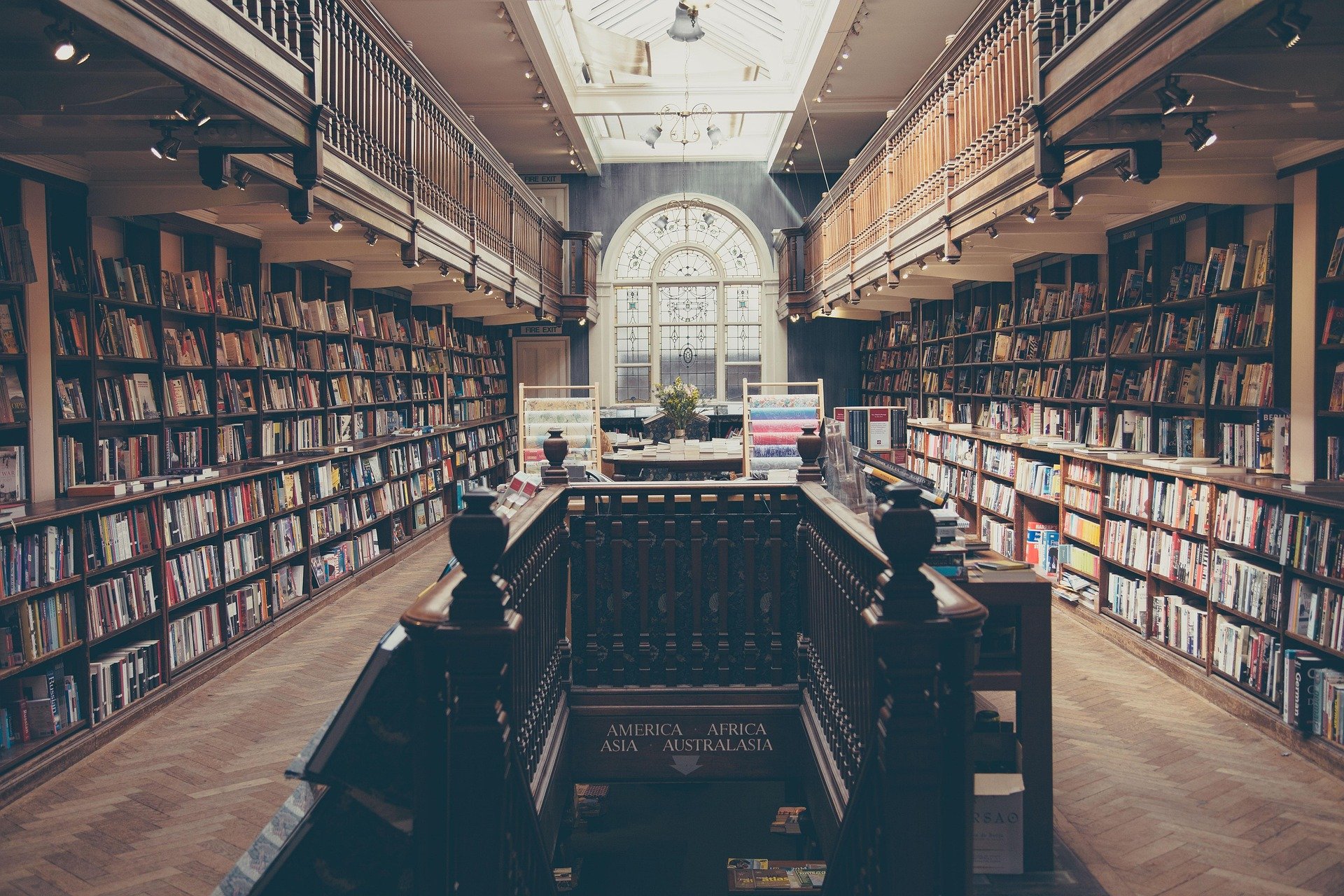 Interior of a historic-style bookstore or library with tall bookshelves lining both sides of the room. The space features a large arched window bringing in natural light, and wooden railings and floors. There are stairs in the foreground leading to a lower level, with labels indicating continents such as America, Africa, Asia, Australasia.