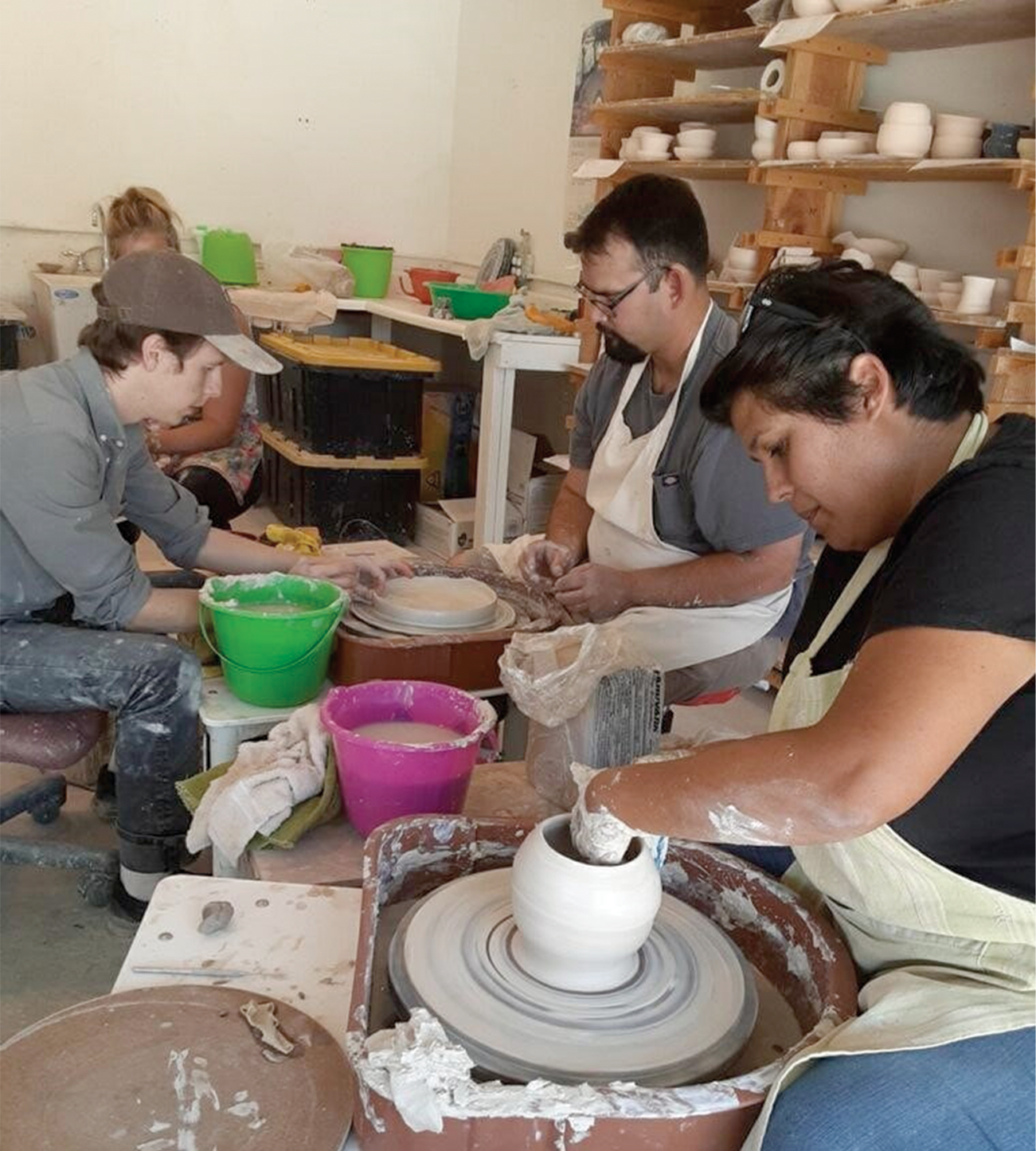 People working on pottery in a ceramics studio with shelves filled with white ceramic bowls and cups.