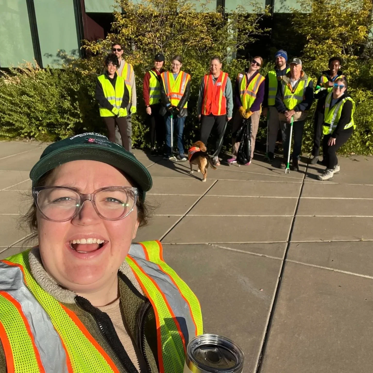 A group of people wearing reflective safety vests posing outdoors in front of bushes and a building with large windows. One person is taking a selfie, with a woman in the foreground smiling and holding a drink. Several others stand behind with a dog on a leash.