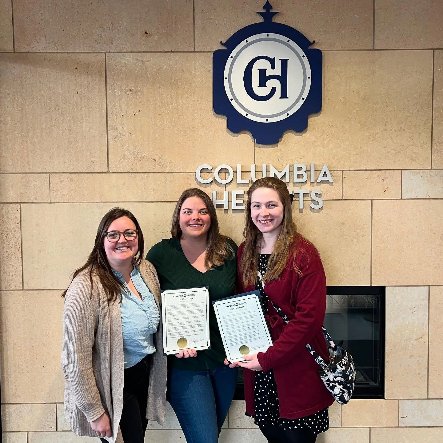 Three women standing in front of a Columbia Heights sign holding certificates. All are smiling and dressed casually.