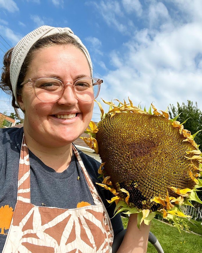 Woman smiling outdoors holding a large sunflower with a clear blue sky behind her.