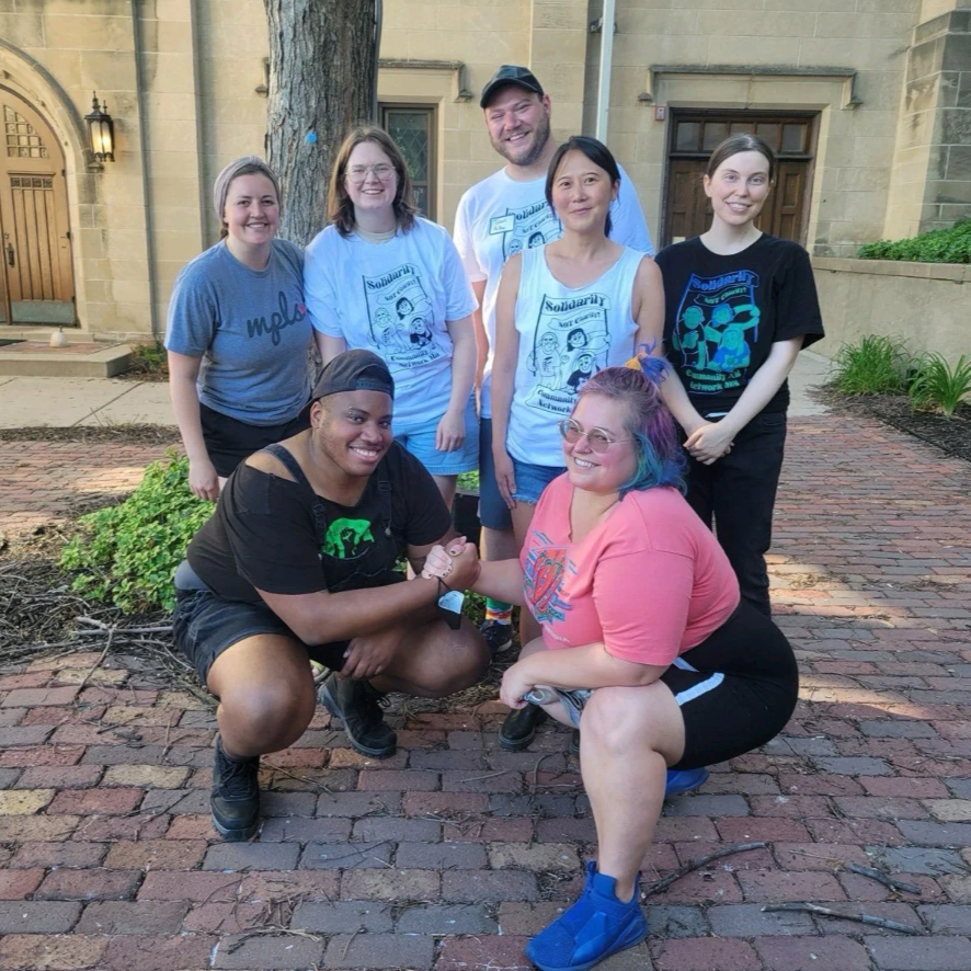 Group of seven diverse people outdoors, smiling and meeting, with some wearing solidarity event shirts, posing on a brick pathway near a stone building with a large tree.