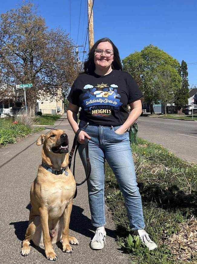 A smiling woman with glasses and long dark hair standing on a sidewalk with her hand in her pocket, holding a leash attached to a happy, tan-colored dog sitting beside her. They are outdoors on a sunny day with trees, houses, and a clear blue sky in the background.