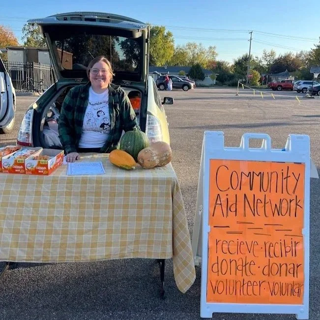 A woman standing behind a table with pumpkins, boxes of food, and a clipboard, set up for a community aid event in a parking lot. There is an orange sign next to the table with the words 'Community Aid Network' and related keywords.