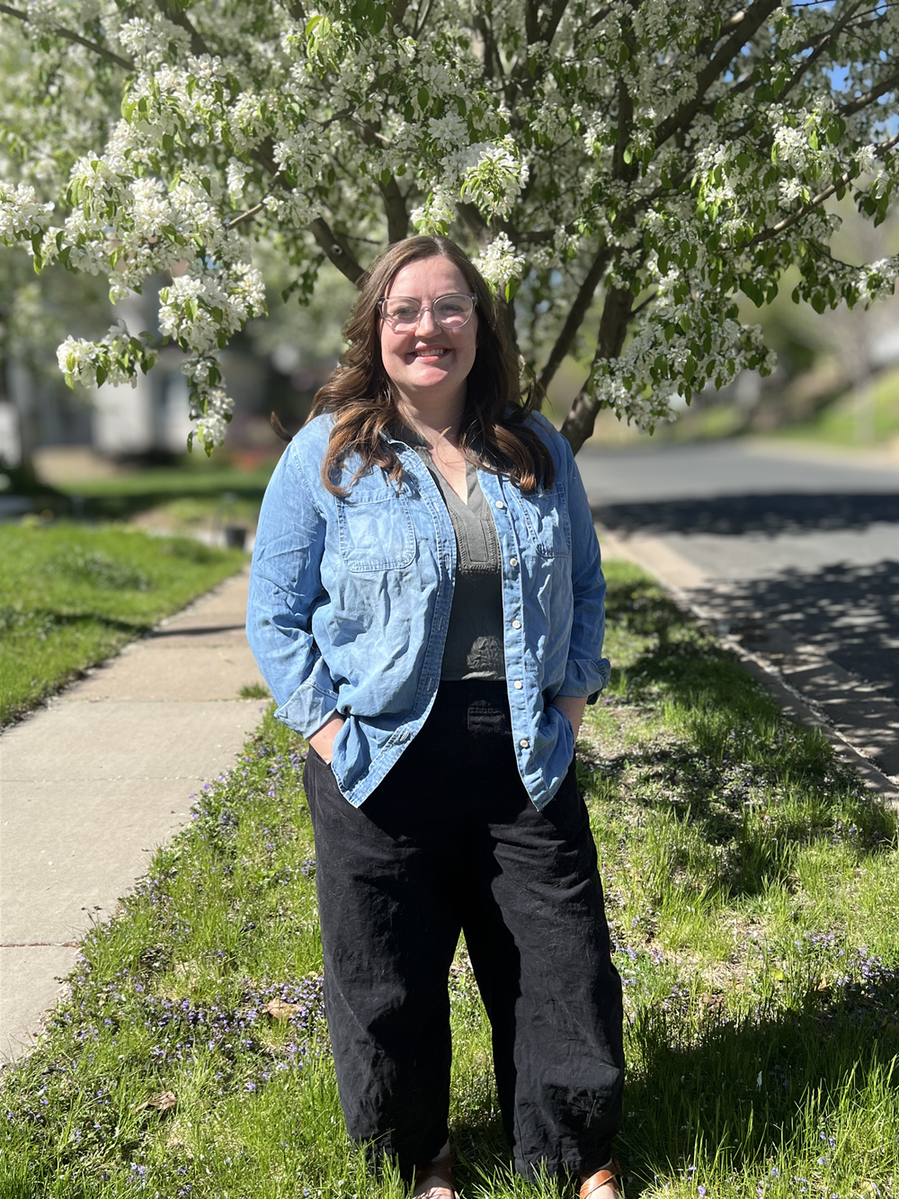 A woman standing on a sidewalk in front of a blooming tree on a sunny day.