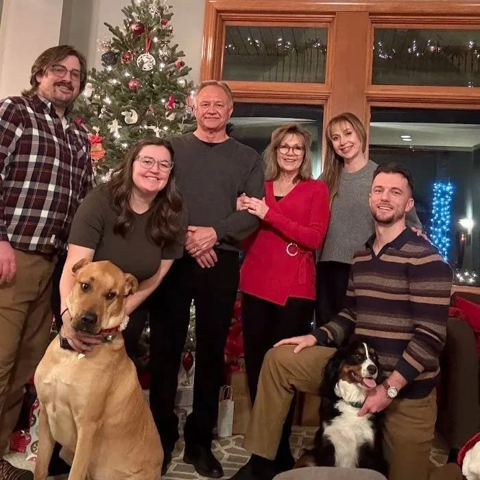 Group of six people and two dogs gathered in a living room decorated for Christmas, with a decorated Christmas tree in the background.