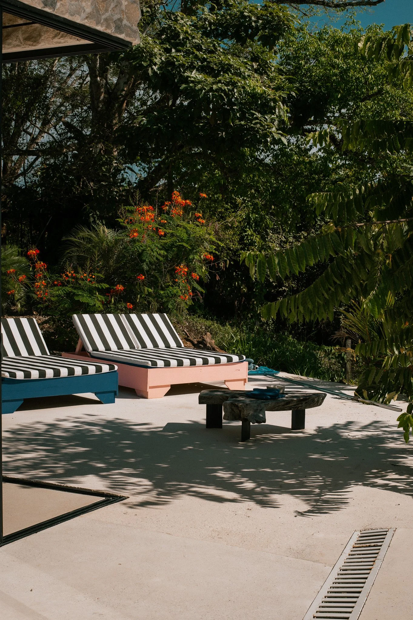 Poolside scene with two lounge chairs with black and white striped cushions, a small wooden table, lush green trees, and vibrant red flowers in the background. Shadows from the trees are cast on the light-colored concrete surface.