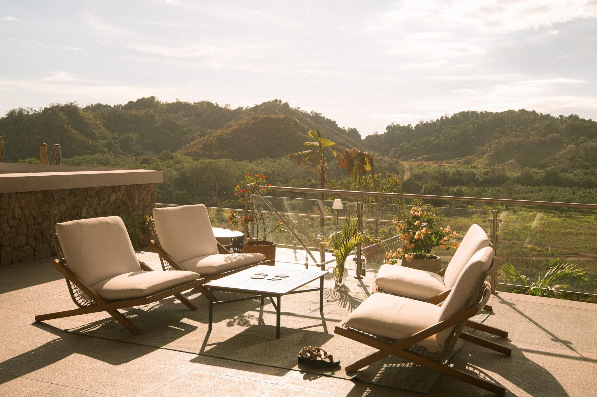 Outdoor patio area with white cushioned lounge chairs and a black metal table, decorated with potted plants and flowers, overlooking green hills and trees in the distance.