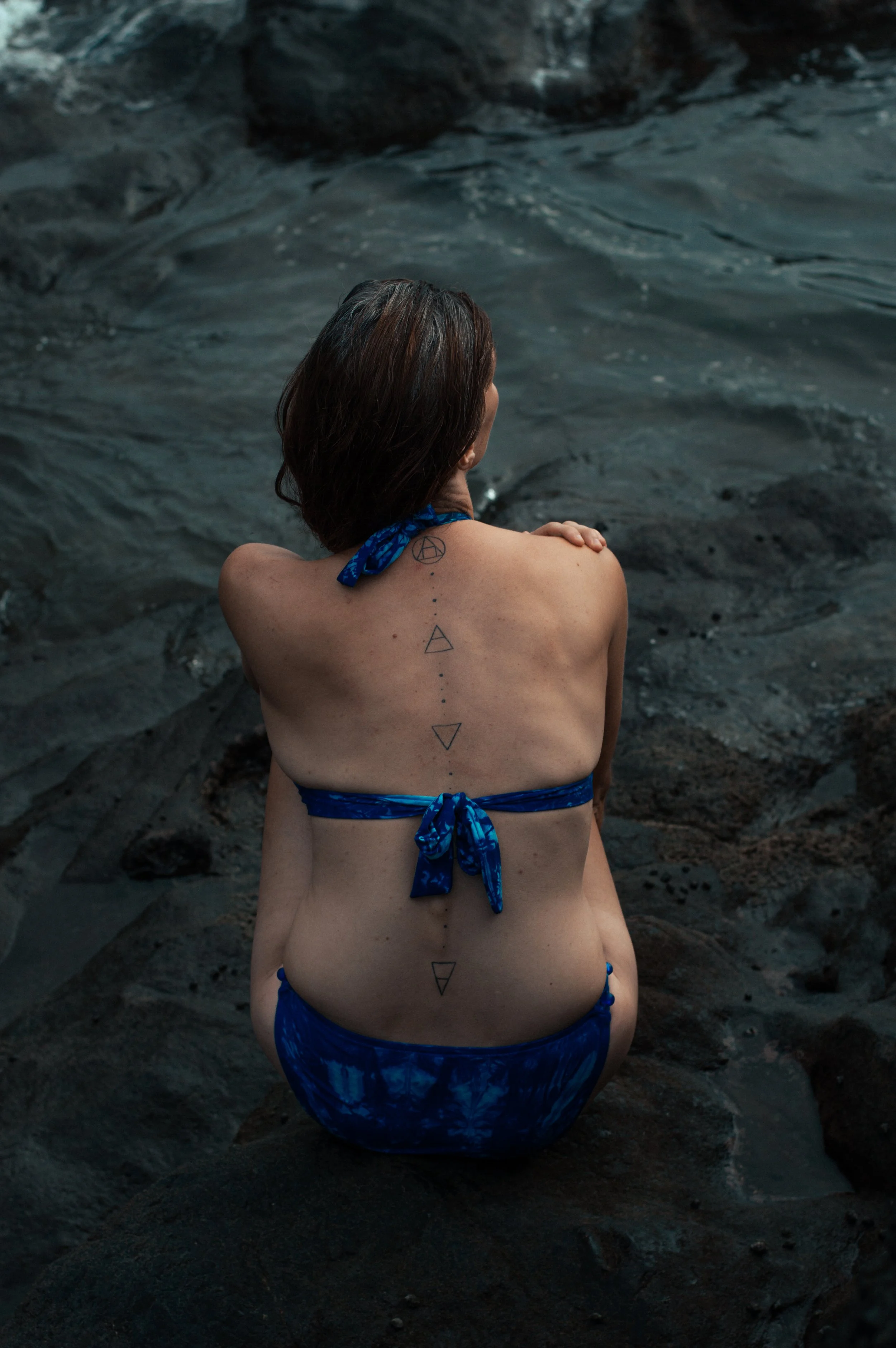 A woman with short brown hair sitting on dark rocks beside water, wearing a blue patterned bikini, with tattoos on her back and shoulders, looking away from the camera.