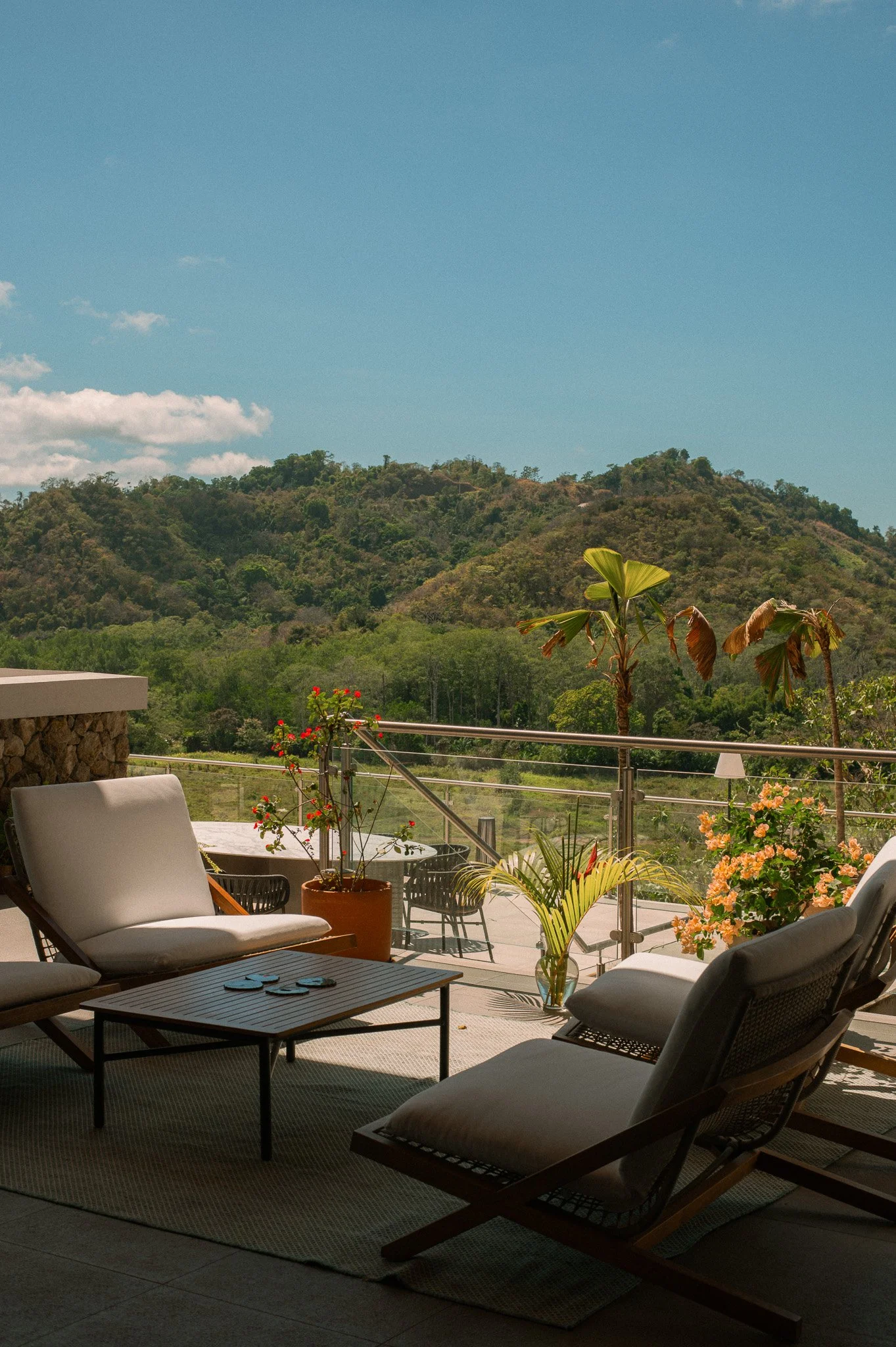 Outdoor patio with white cushioned chairs, a small table, and potted plants, overlooking lush green hills and a blue sky with a few clouds.