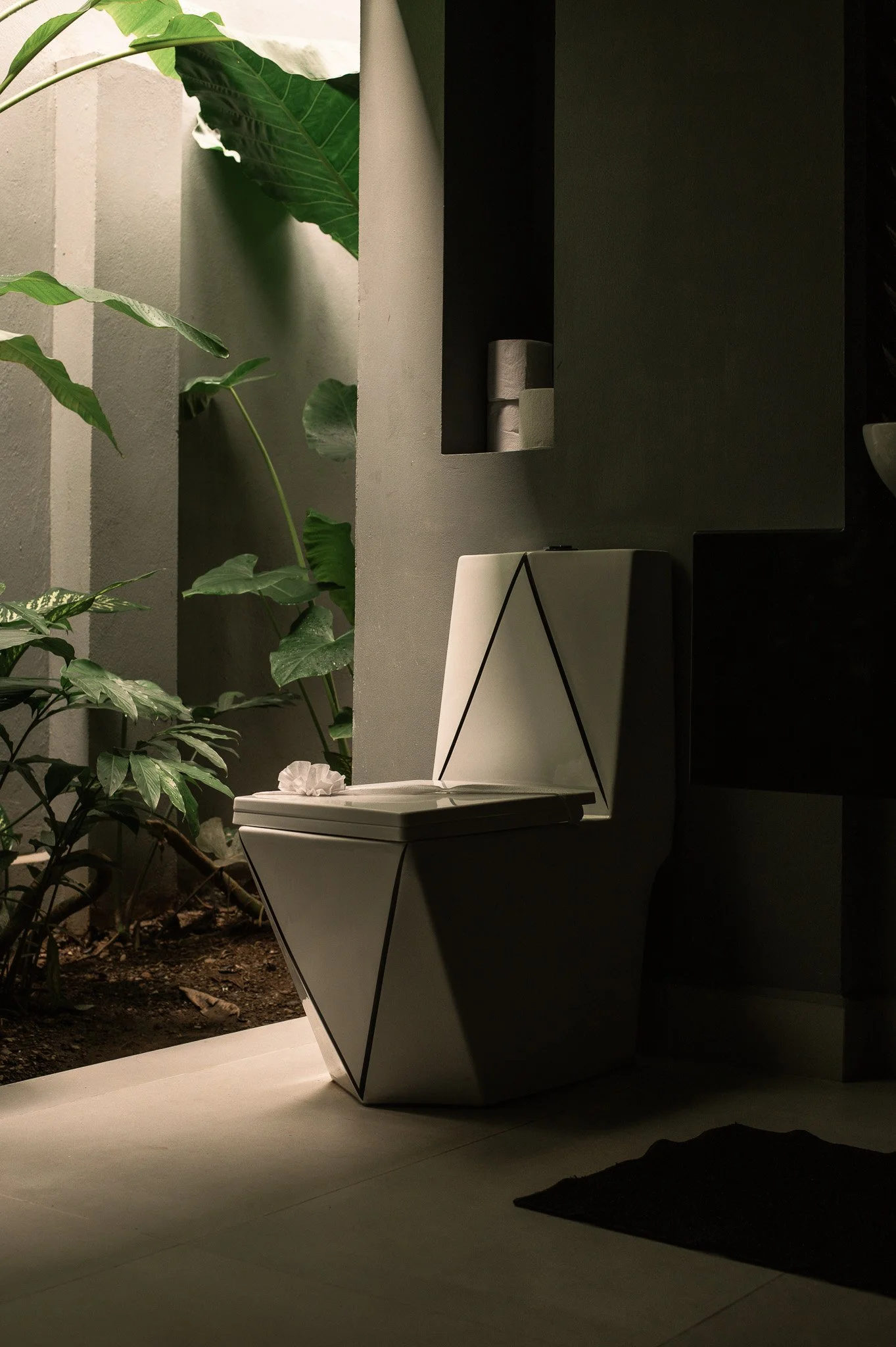 Indoor scene with a modern, geometric white toilet on a light-colored floor, situated near a garden with green leafy plants. The background features a textured wall and a black geometric structure with small stacked rolls or containers. The lighting 