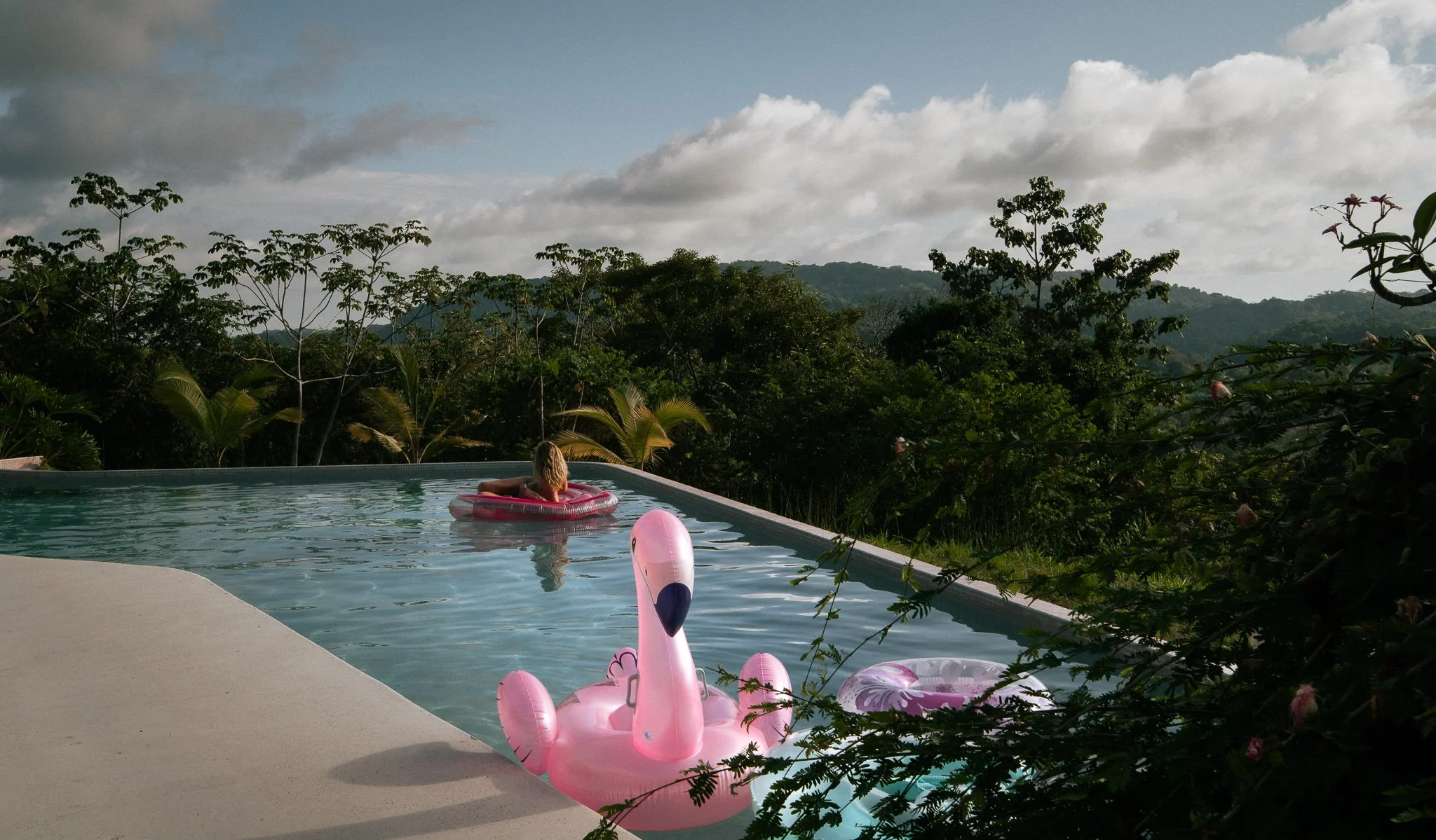 A girl with blonde hair floating on a pink inner tube in an outdoor swimming pool, with a view of lush green trees and hills in the background, under a partly cloudy sky.