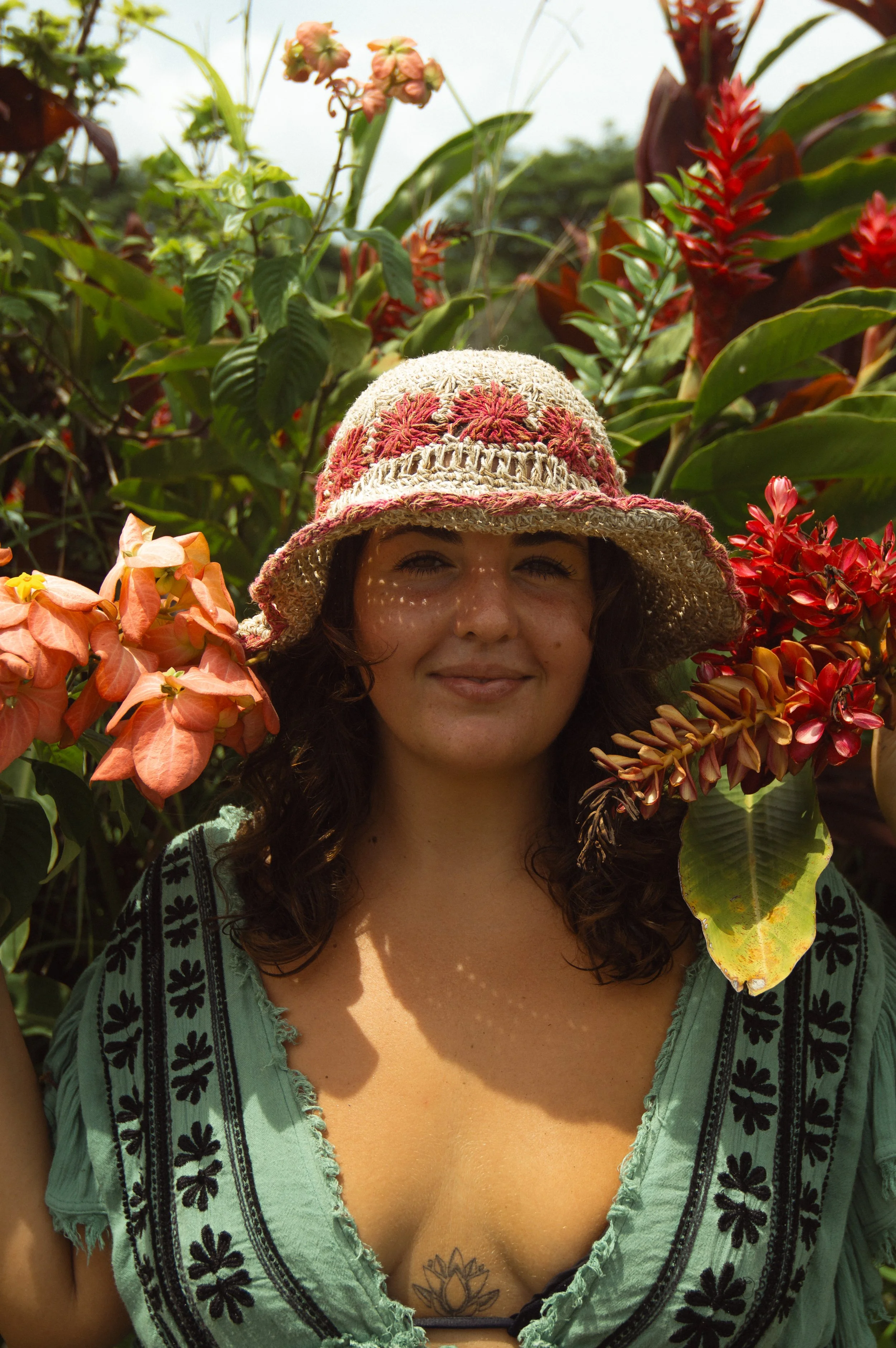 Woman with curly hair wearing a wide-brimmed straw hat and a green top with black floral embroidery, standing among lush green and red plants in a garden.