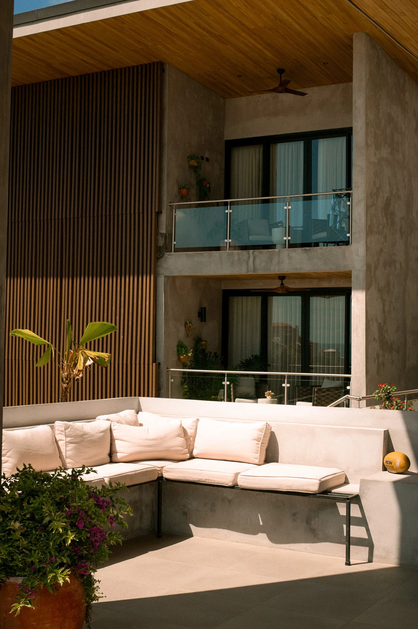 An outdoor patio area with a white curved sectional sofa and cushions, potted plants, a wooden slatted wall, glass railing balcony with interior view of a room, ceiling fan, and a volleyball on the ground.