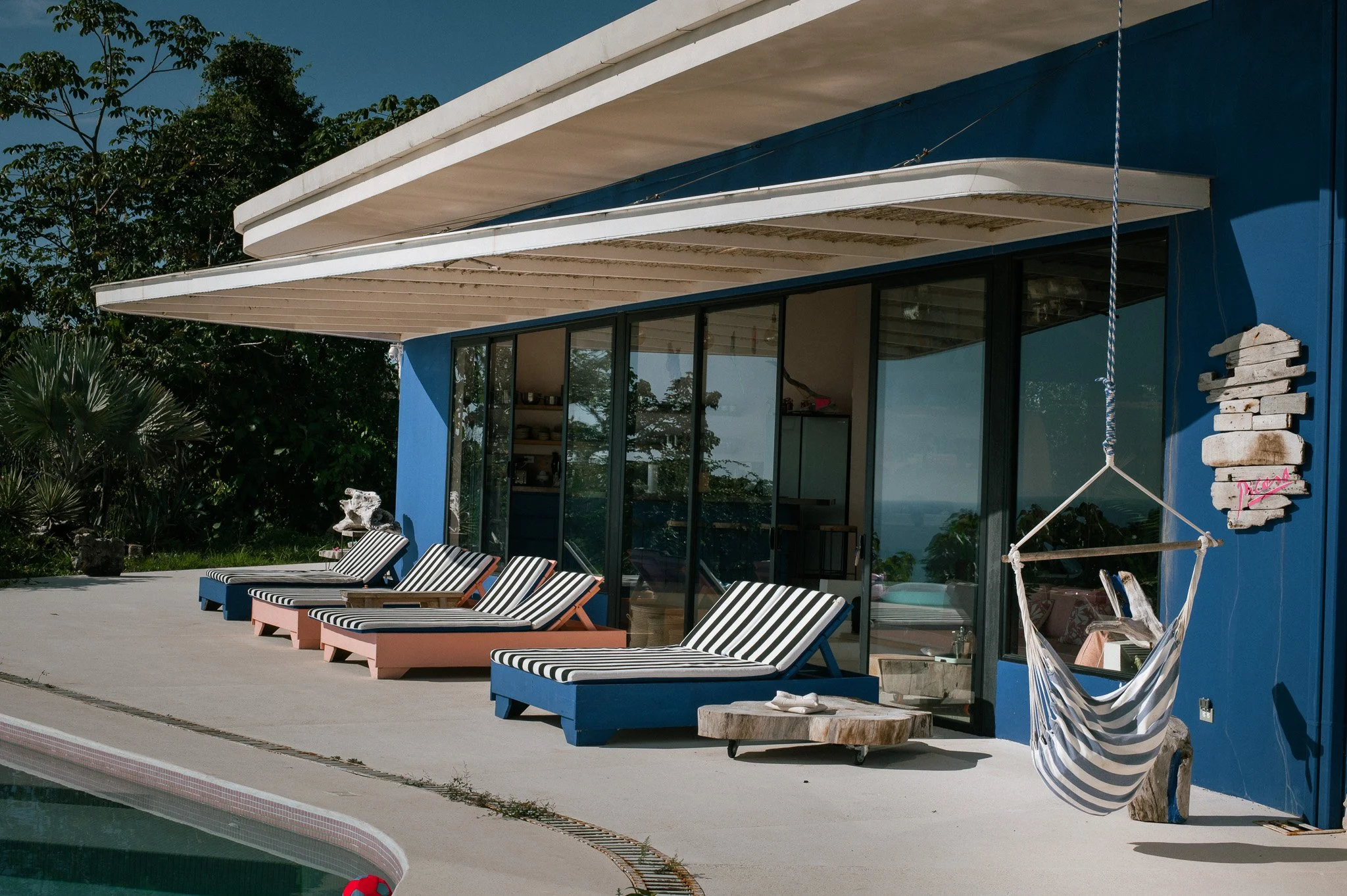 A modern house with a blue exterior and large glass doors, outdoor striped lounge chairs, a hammock, and a poolside patio with lush greenery in the background.