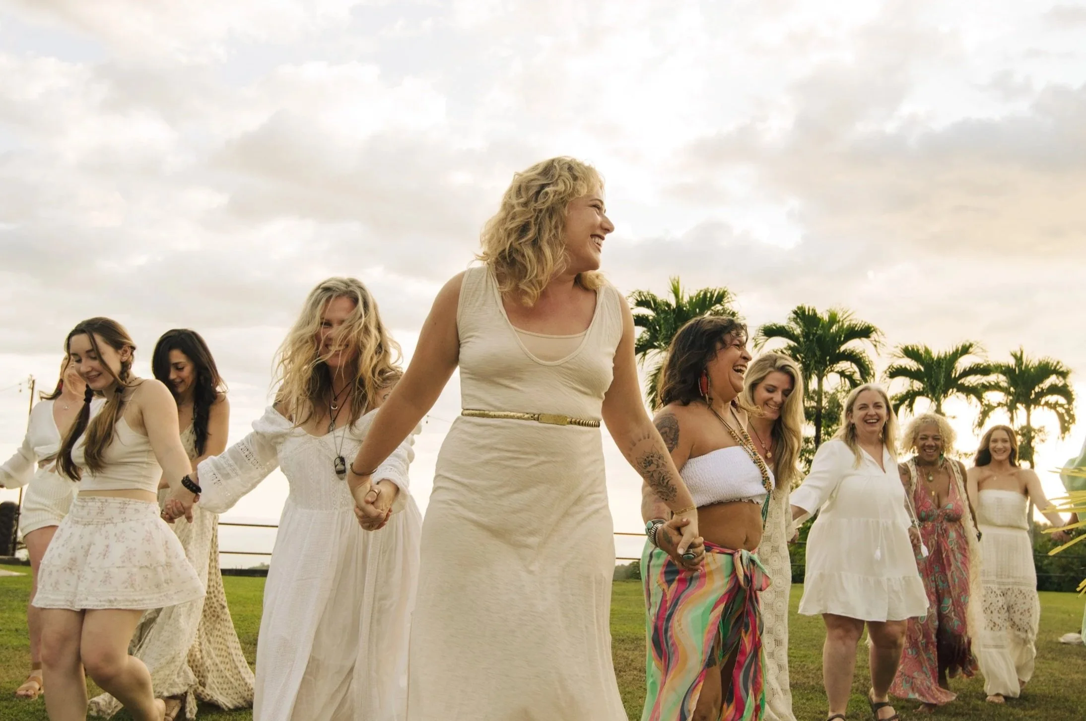 A group of women dancing outdoors on a grassy field under a cloudy sky, holding hands and smiling. The women are dressed in bohemian style clothing with natural and colorful patterns.