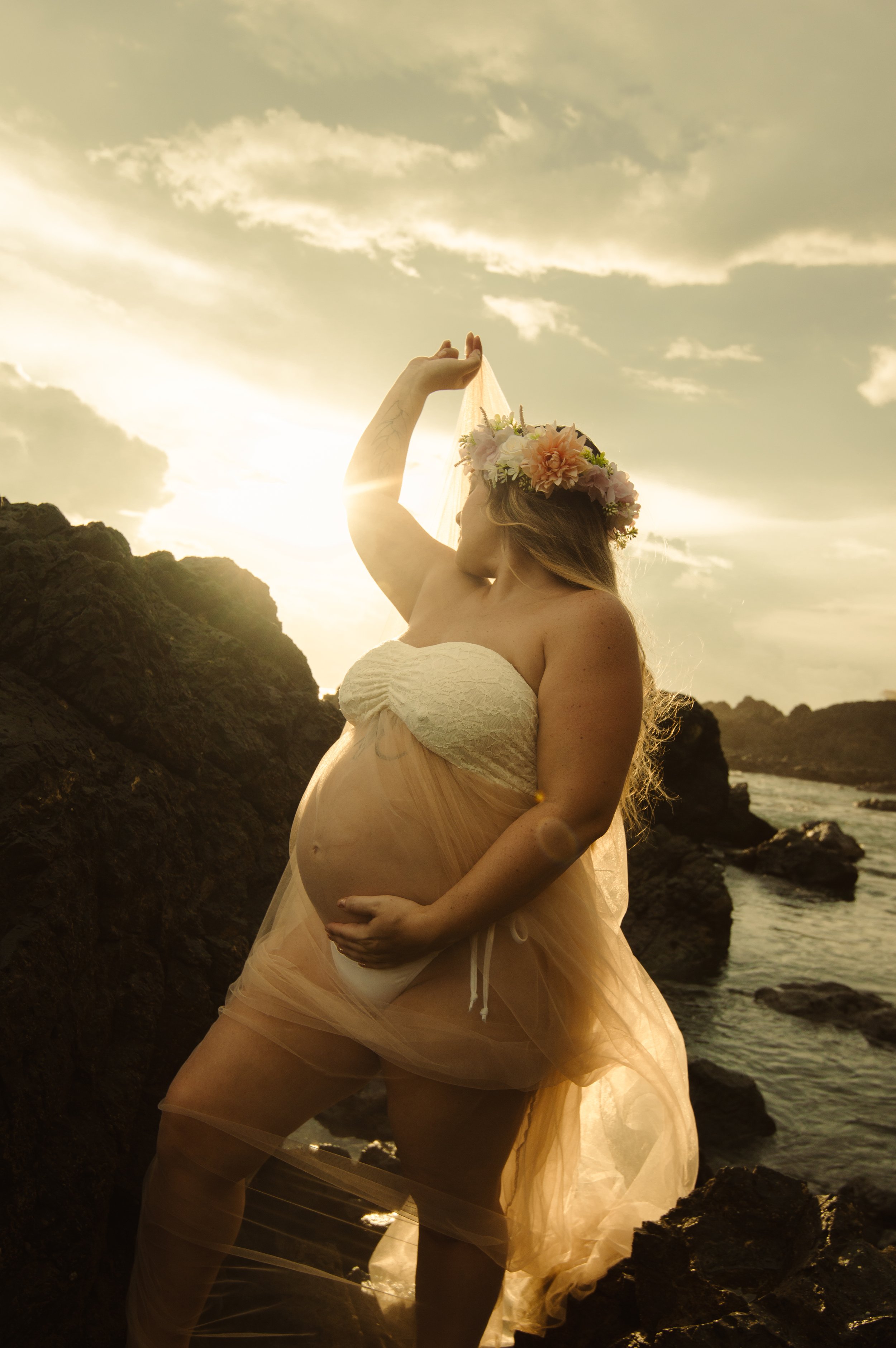 A pregnant woman wearing a floral crown stands near rocks and the ocean at sunset, holding her belly and raising her hand towards the sky.